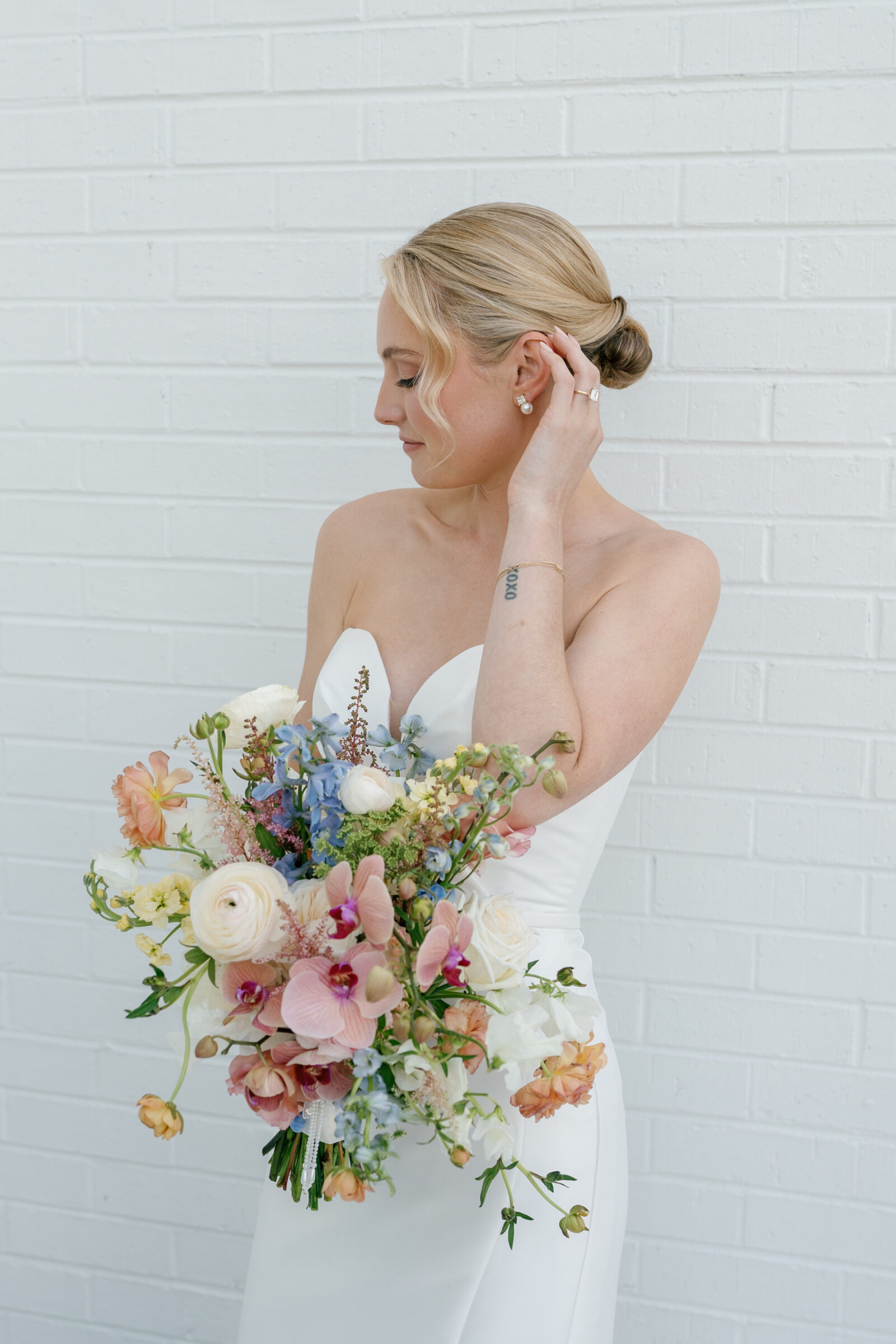Northfield Estate Wedding - Richmond VA | Profile view of a woman in a strapless white gown adjusting a pearl drop earring as she holds a lush bouquet of pastel orchids, ranunculus, delphinium, and greenery against a white brick wall.