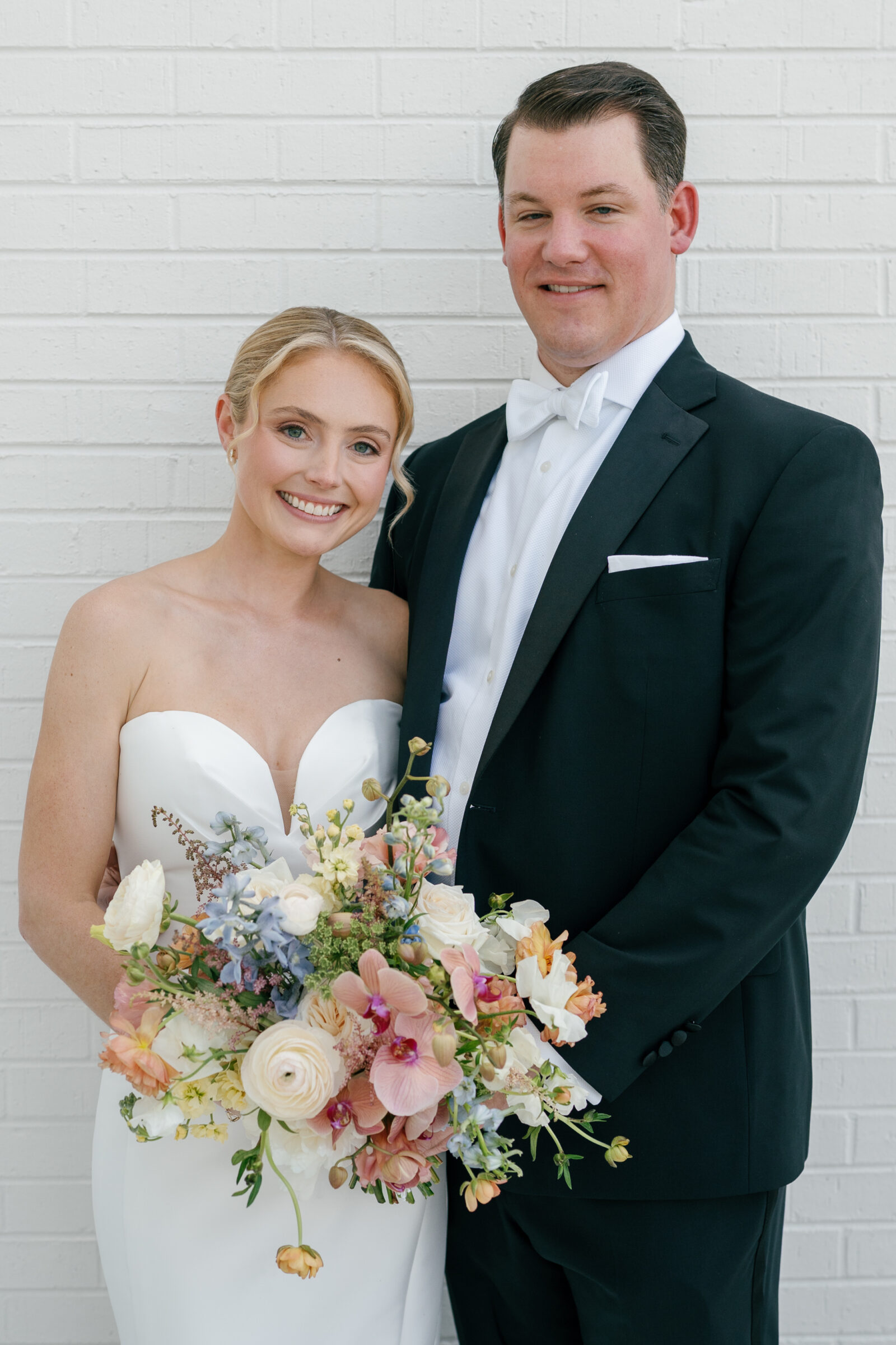 Portrait of a bride and groom standing side by side against a white brick wall; the bride wears a strapless satin gown and holds a lush spring bouquet of pink orchids, creamy ranunculus, and blue delphinium, while the groom stands beside her in a black tuxedo with a white bow tie.