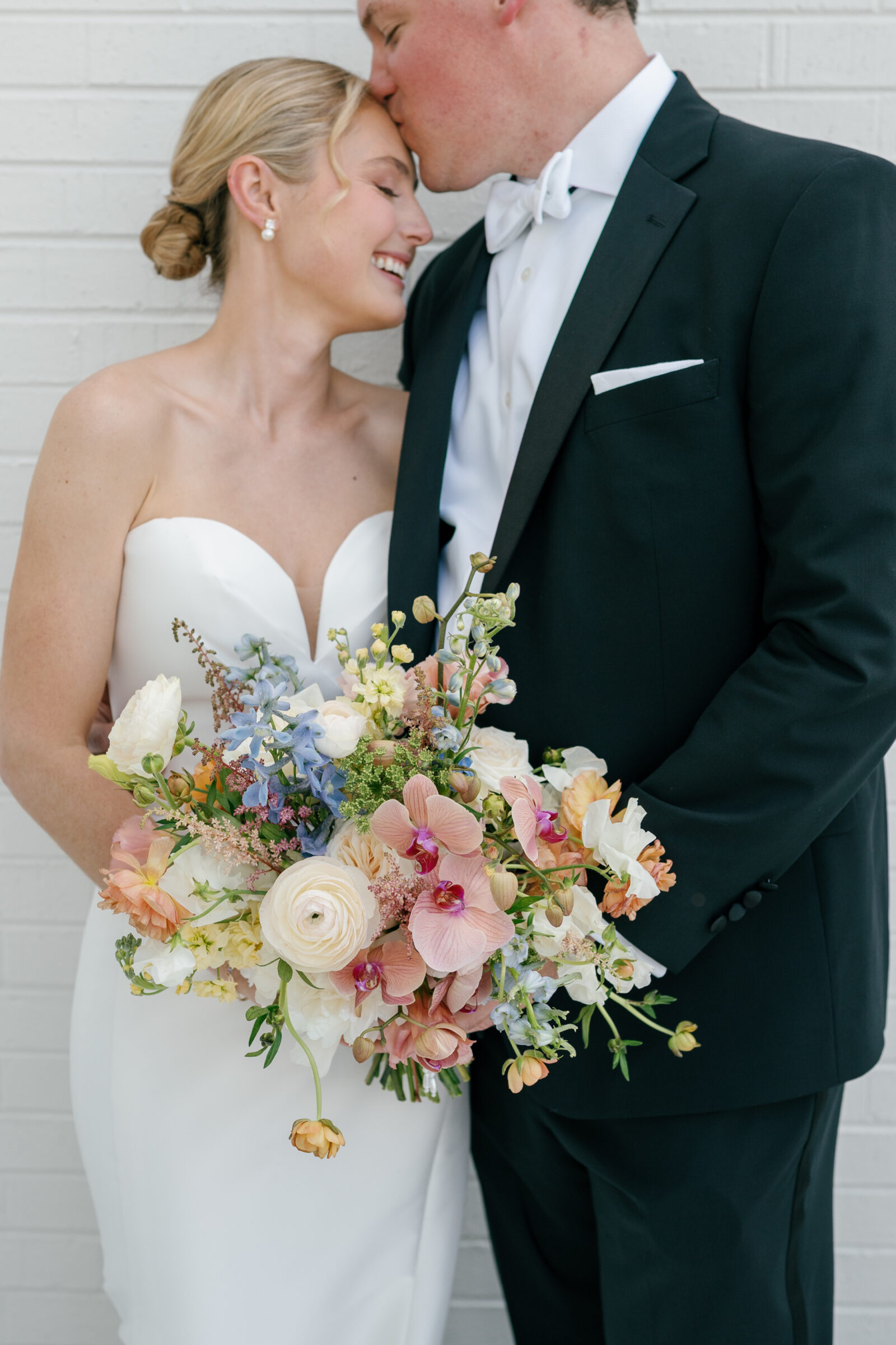 Close-up of the bride and groom standing against a white brick wall, the groom gently kissing the bride’s forehead as they hold a lush spring bouquet filled with pink orchids, creamy ranunculus, pale blue delphinium, and trailing greenery.
