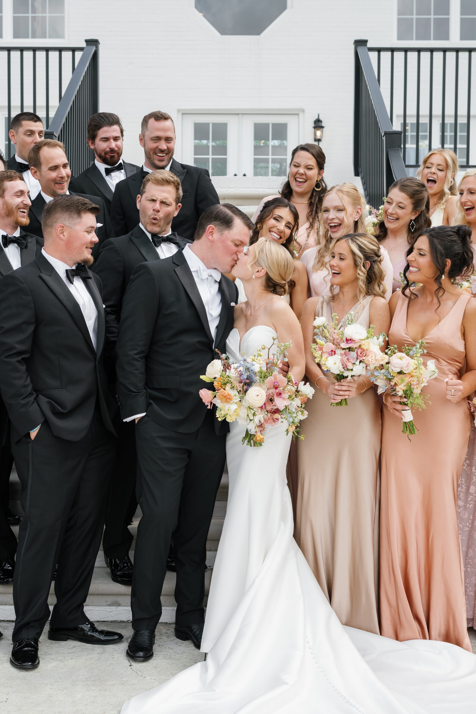 Large wedding party portrait on outdoor stone steps: the bride and groom share a kiss at center, surrounded by groomsmen in black tuxedos and bridesmaids in mixed pastel gowns, all laughing and cheering beneath a white brick facade.
