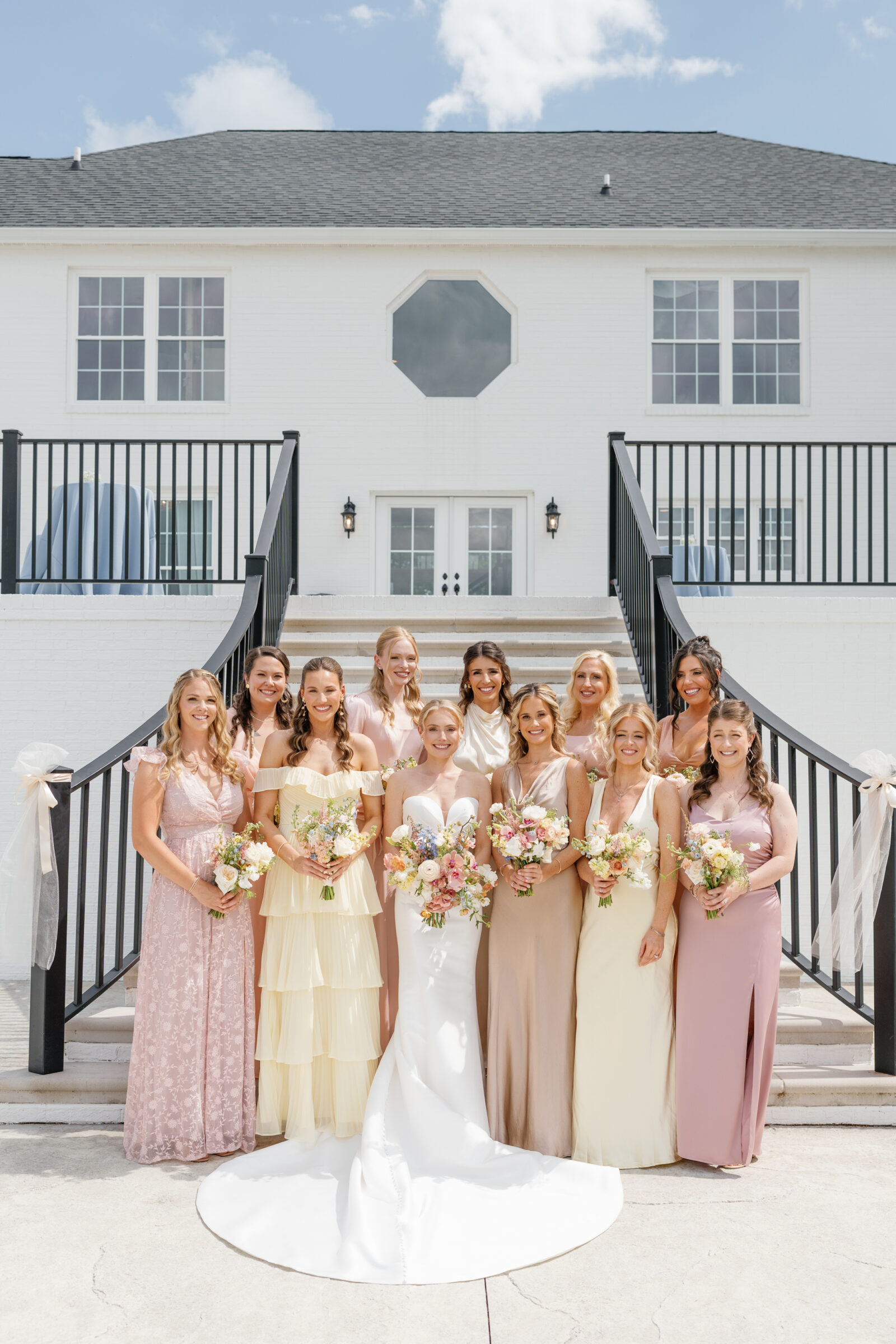 Northfield Estate Wedding - Richmond VA | Wide group portrait of the bride and her bridesmaids posed on stone steps in front of the estate’s white brick façade: the bride wears a strapless white satin gown and stands at center, while ten bridesmaids flank her in mixed pastel dresses—soft pink lace, buttery yellow tiers, champagne satin, blush—and each holds a hand‑tied spring bouquet.