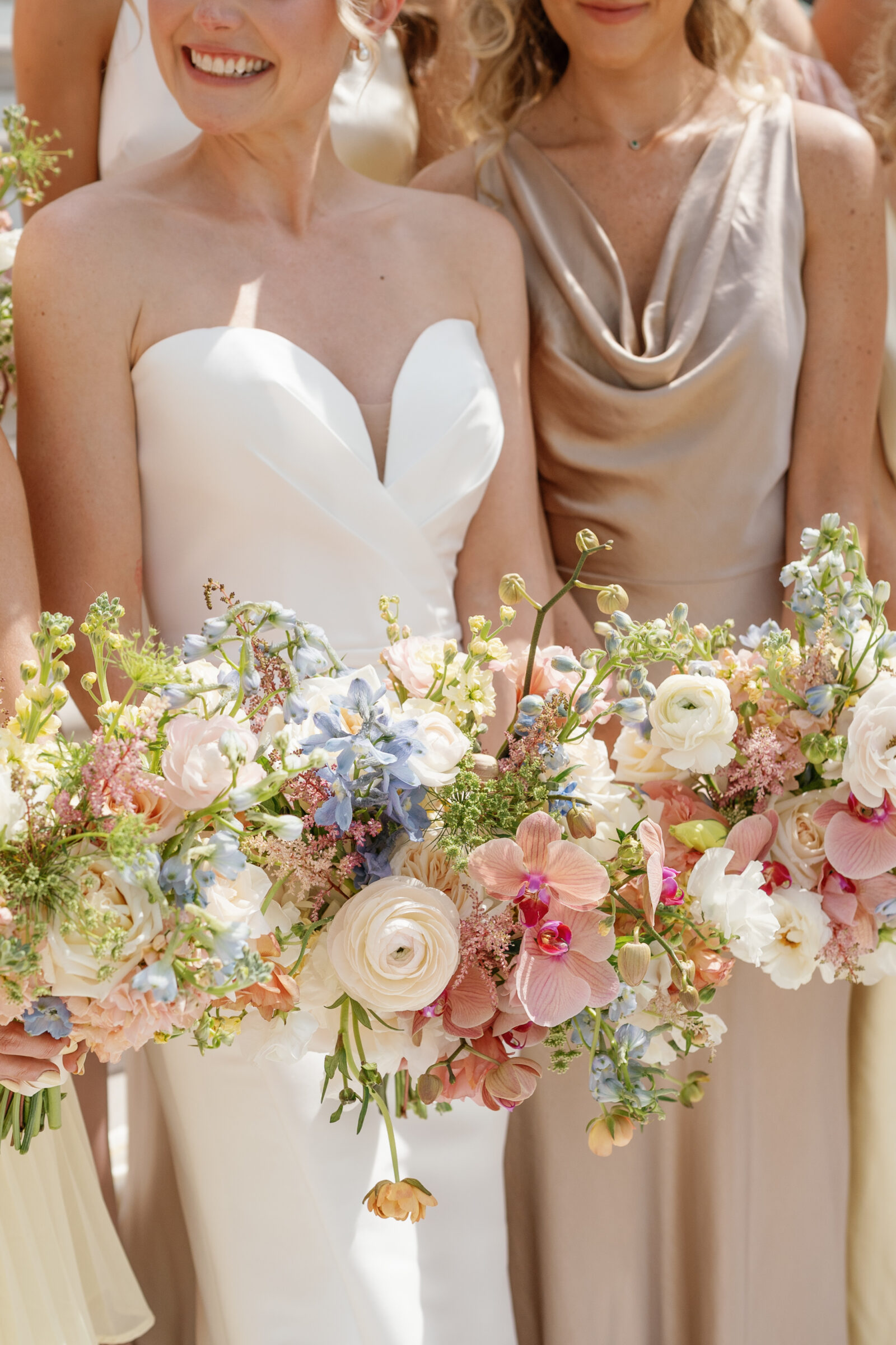 Close-up of the bouquets held by the bride and her bridesmaids, showcasing lush arrangements of pink orchids, creamy ranunculus, pale blue delphinium, peach ranunculus, and delicate astilbe, all accented with trailing greenery against their pastel-toned dresses.