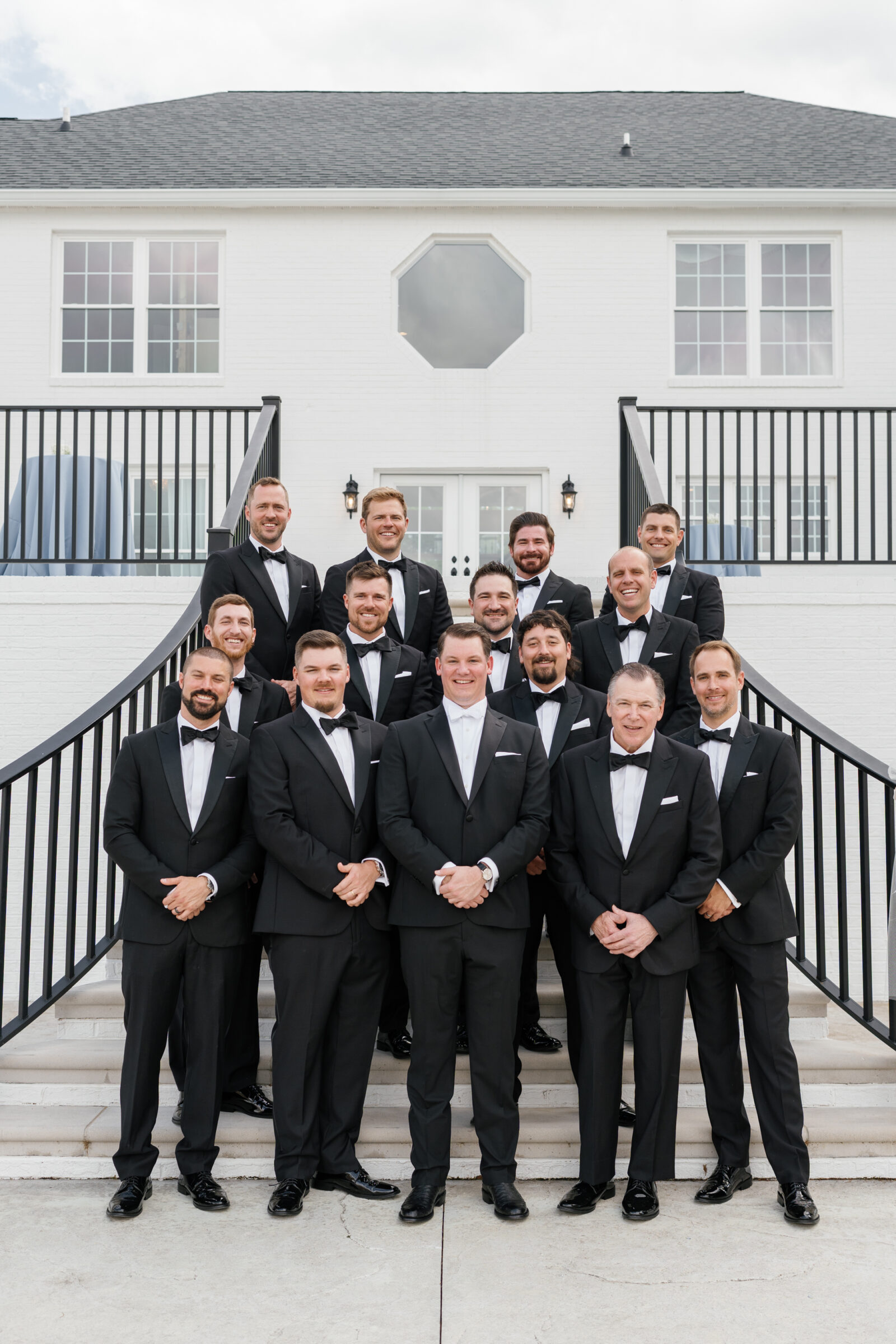 Wide portrait of the groom surrounded by his groomsmen on stone steps in front of a white‑brick estate entrance; all wear black tuxedos and bow ties, standing in neat rows beneath black iron railings.