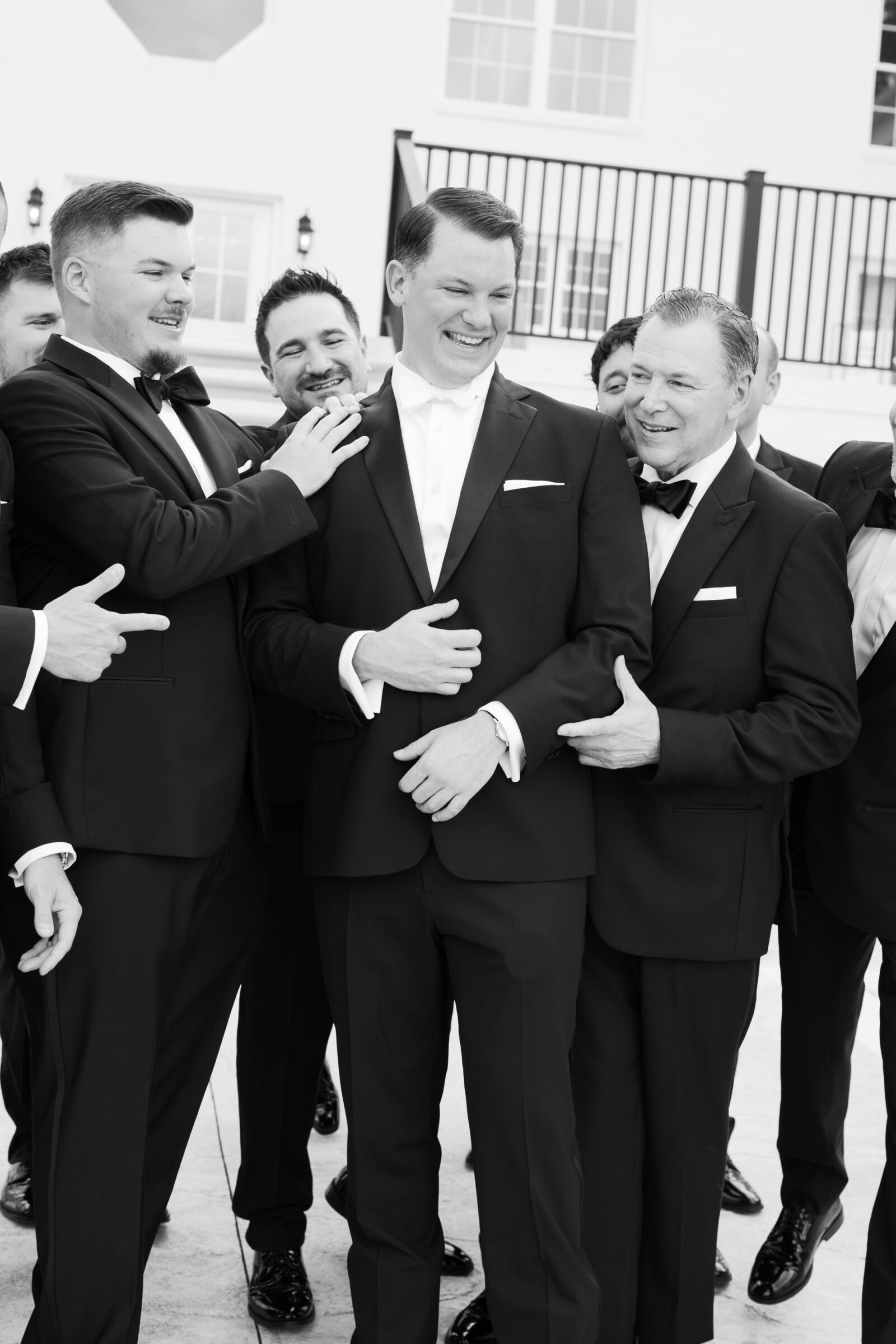 Black-and-white close‑up of the groom laughing as his groomsmen gather around him on the steps, their hands resting on his shoulders and pointing playfully, all dressed in classic tuxedos.