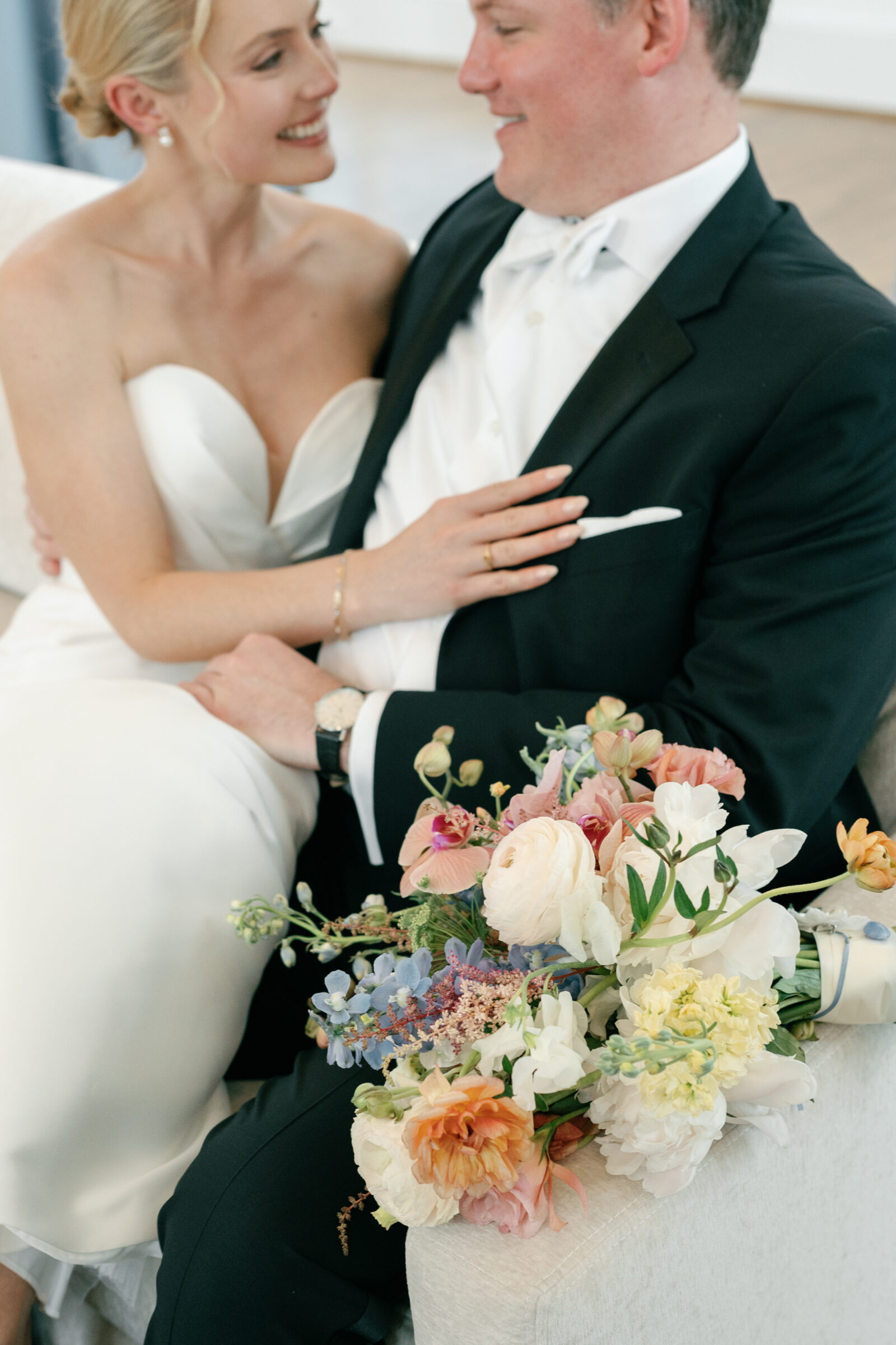 A bride and groom seated together on a light-colored sofa, smiling at each other as she rests her arm across his chest with a lush spring bouquet of pastel blooms draped over the sofa arm in the foreground.