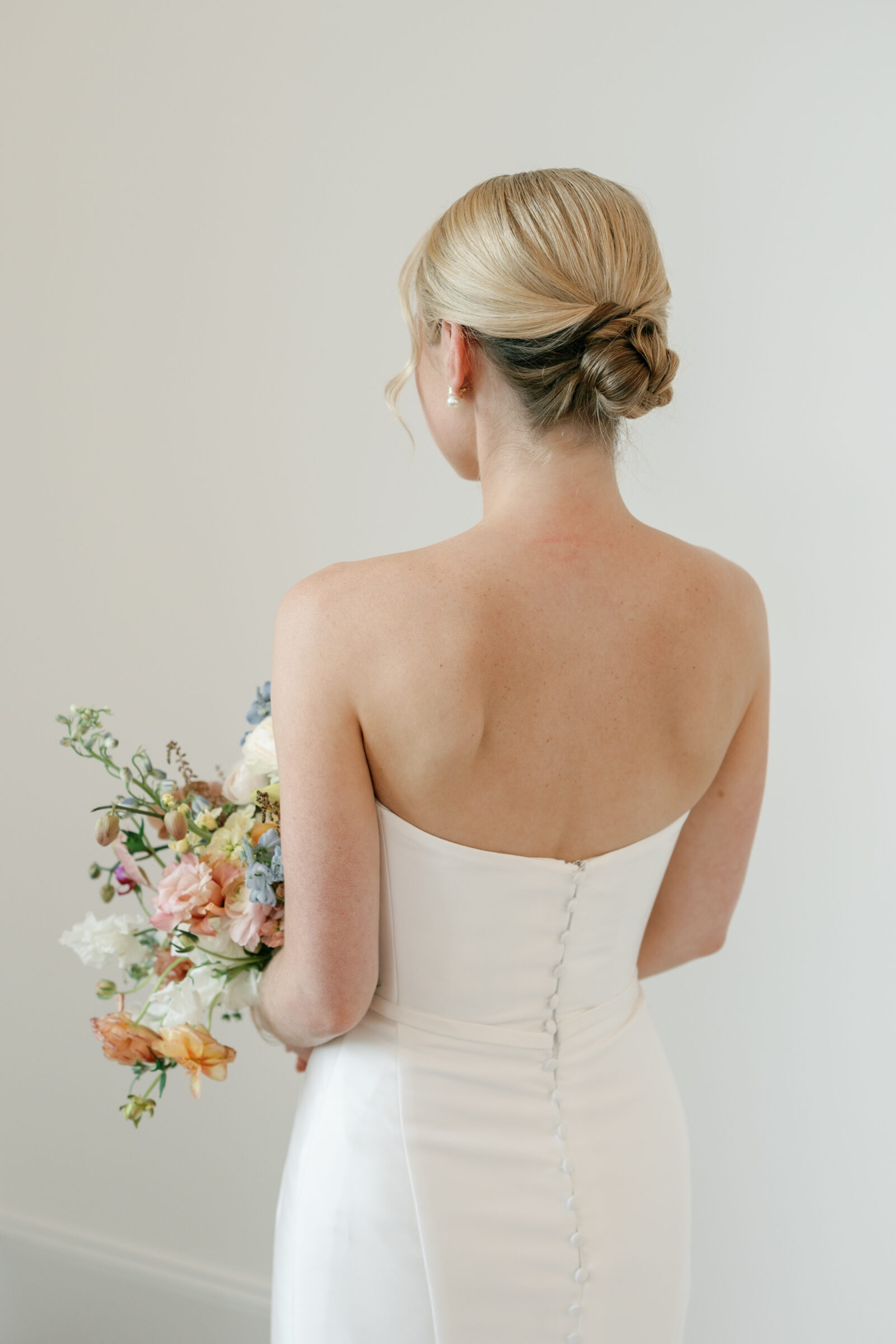 The bride seen from behind, standing against a neutral wall in her strapless satin gown with a row of fabric‑covered buttons down the back, holding a loose, garden‑style bouquet of pastel orchids, ranunculus, delphinium, and greenery.
