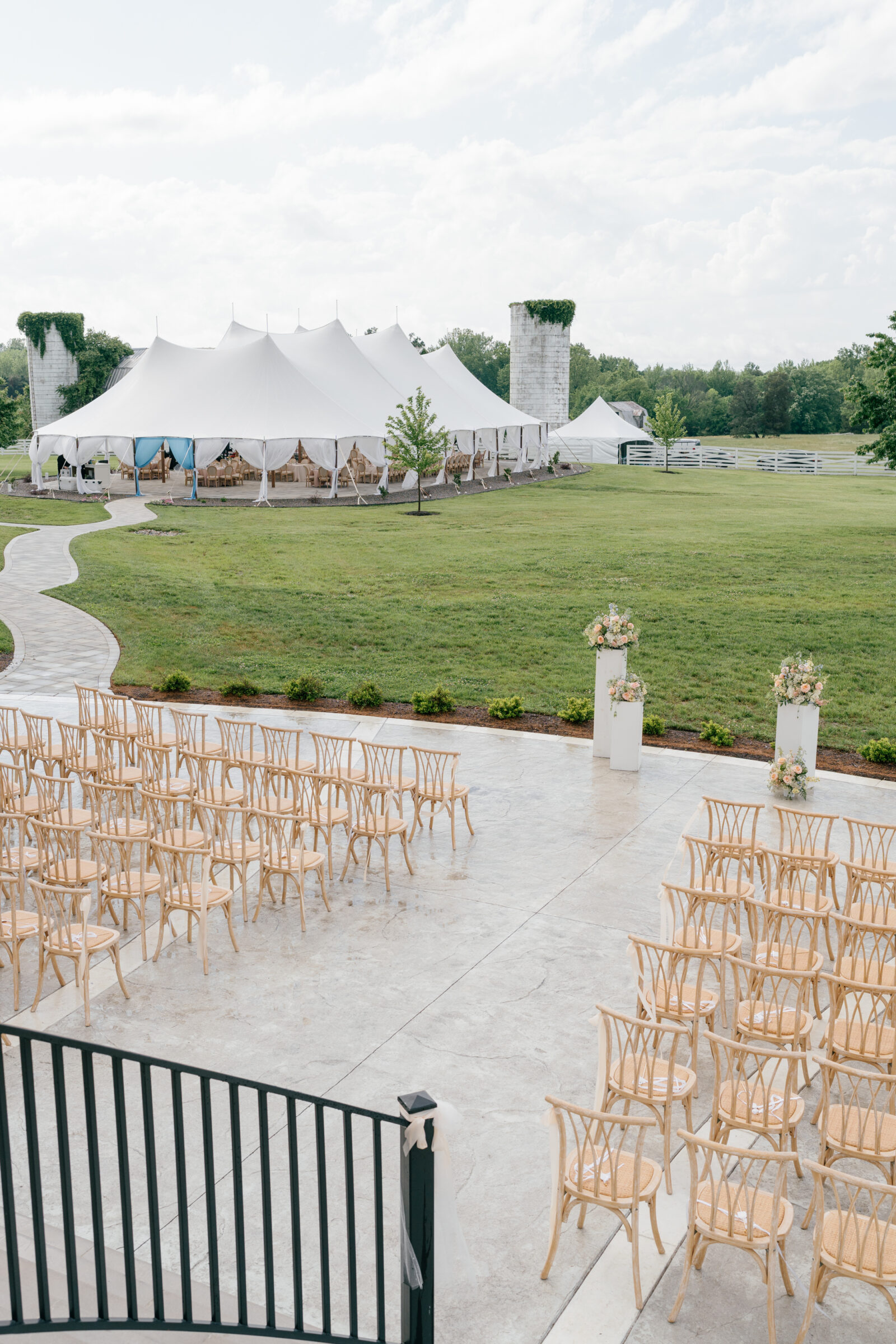 Northfield Estate Wedding - Richmond VA | Wide view of the outdoor wedding grounds at Northfield Estate, featuring rows of natural wood cross‑back chairs set on a stone terrace facing three white pedestal arrangements of pastel blooms, with a large white sailcloth reception tent, verdant lawn, and ivy‑topped silos under a partly cloudy sky.