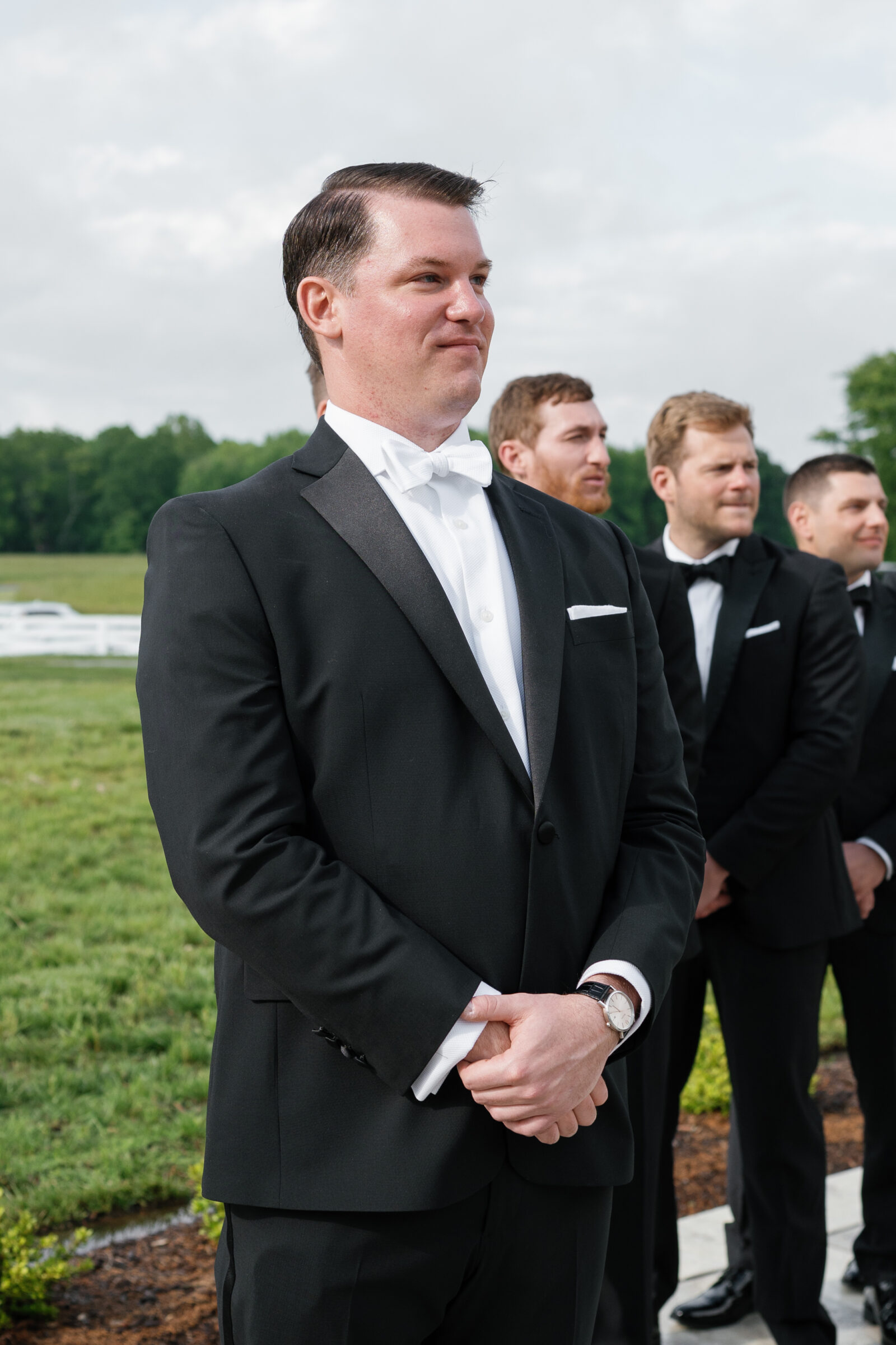 Groom in a black tuxedo and white bow tie stands at the edge of a grassy ceremony lawn, hands clasped in front of him, while a line of groomsmen in matching tuxedos waits behind him beneath a partly cloudy sky.