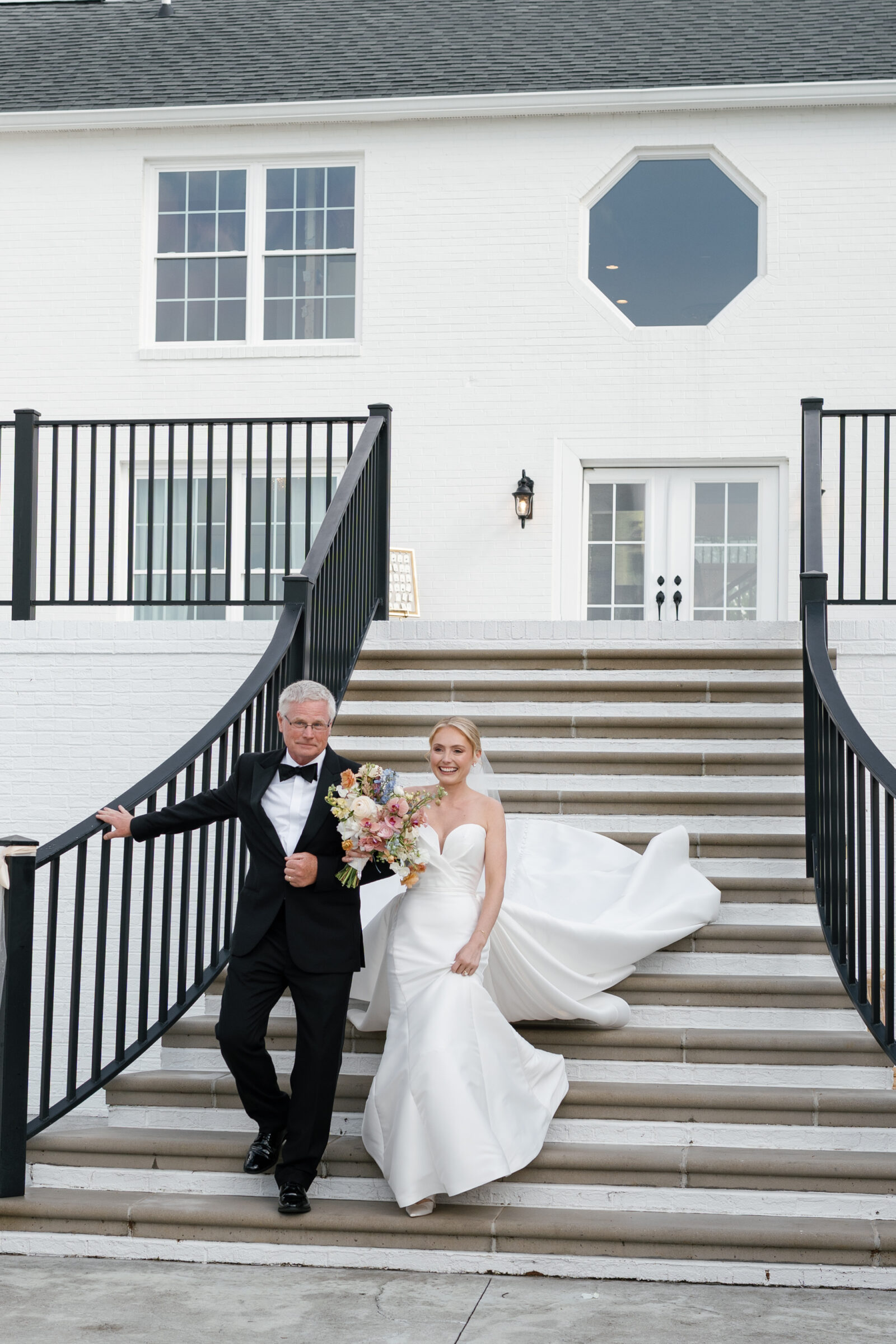 Bride and her father descend wide white‑brick steps of the estate’s entrance; he in a black tuxedo steadies himself on the iron railing as she in a strapless satin gown holds a spring bouquet and lets her train flow behind her.