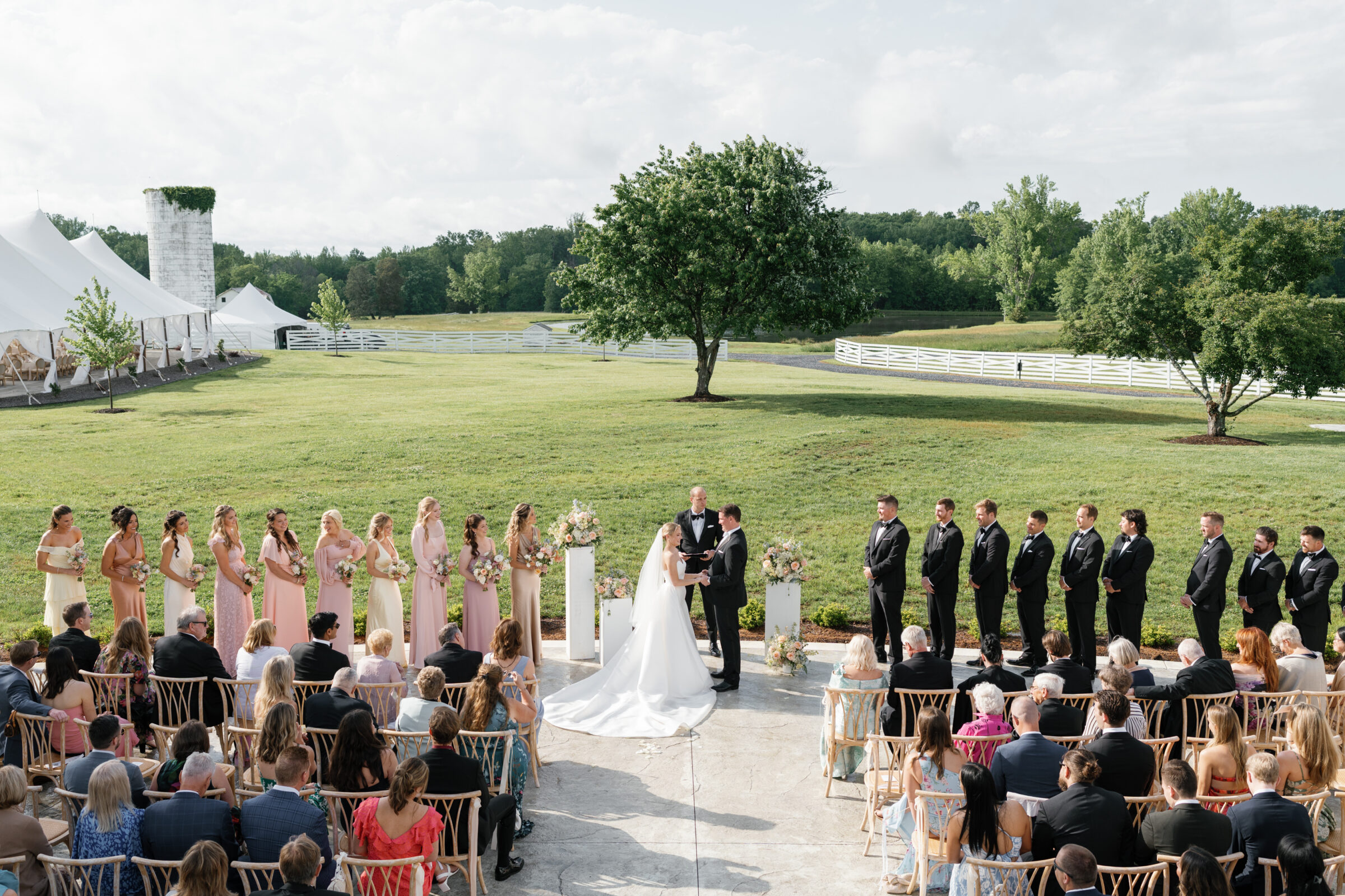 Wide-angle shot of an outdoor ceremony on a concrete terrace in front of a green lawn at Northfield Estate. The couple stands at the center beneath a simple arch, holding hands as the officiant faces them. To the left, a line of bridesmaids in soft pastel dresses holds bouquets; to the right, groomsmen in black tuxedos stand in a matching row. Guests are seated in natural‑wood chairs in front, and a sailcloth reception tent and a white silo peek out on the far left under a partly cloudy sky.