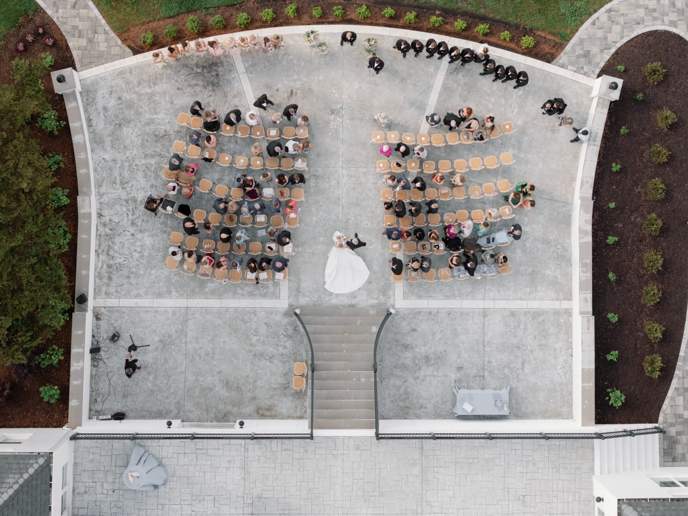 Aerial view of the ceremony terrace at Northfield Estate: two blocks of natural‑wood chairs on a concrete patio flank a central aisle leading up to a short flight of white brick steps. The bride and groom stand together at the top of the aisle facing guests seated in the rows below, with the bridal party lined along the perimeter and lush landscaping edging the stone terrace.