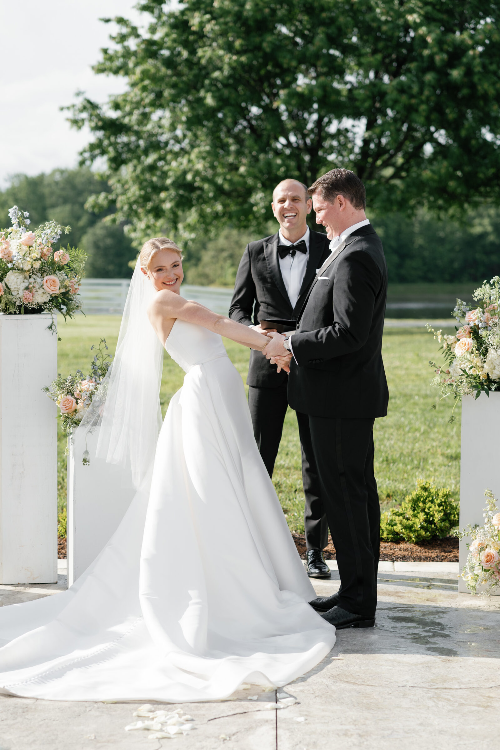A playful moment during the outdoor ceremony: the bride in a strapless white gown leans back with a big smile as she grips the groom’s hand, the officiant stands behind them laughing. Tall white pedestals on either side hold lush arrangements of peach roses, white blooms, and greenery, and a tree and pond fill the sunlit background.