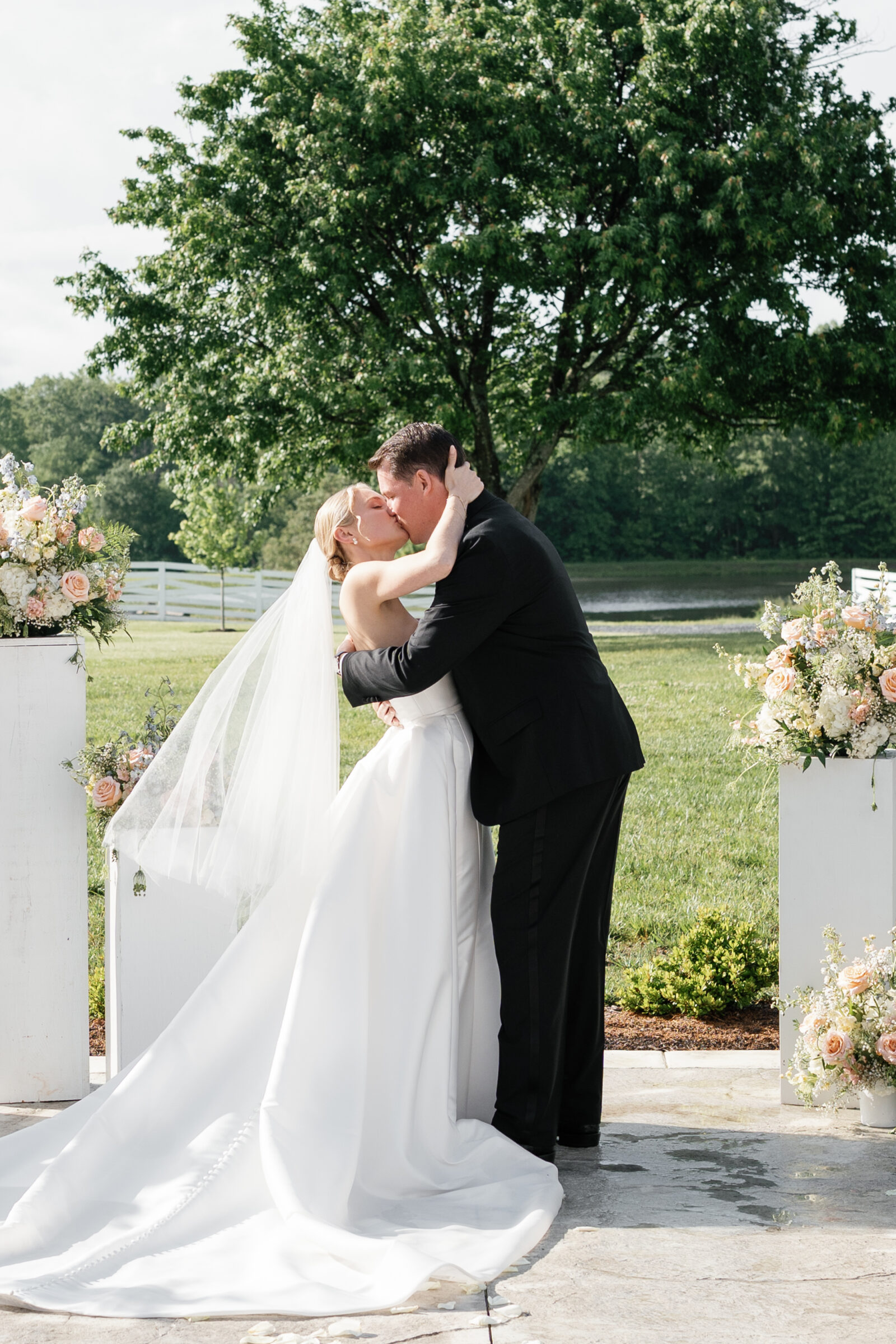 The newly married couple’s first kiss: beneath a leafy tree by a green lawn and white fence, the bride and groom embrace tightly as they share a kiss. The bride’s veil drapes down her back, and matching floral pedestals frame them on either side.