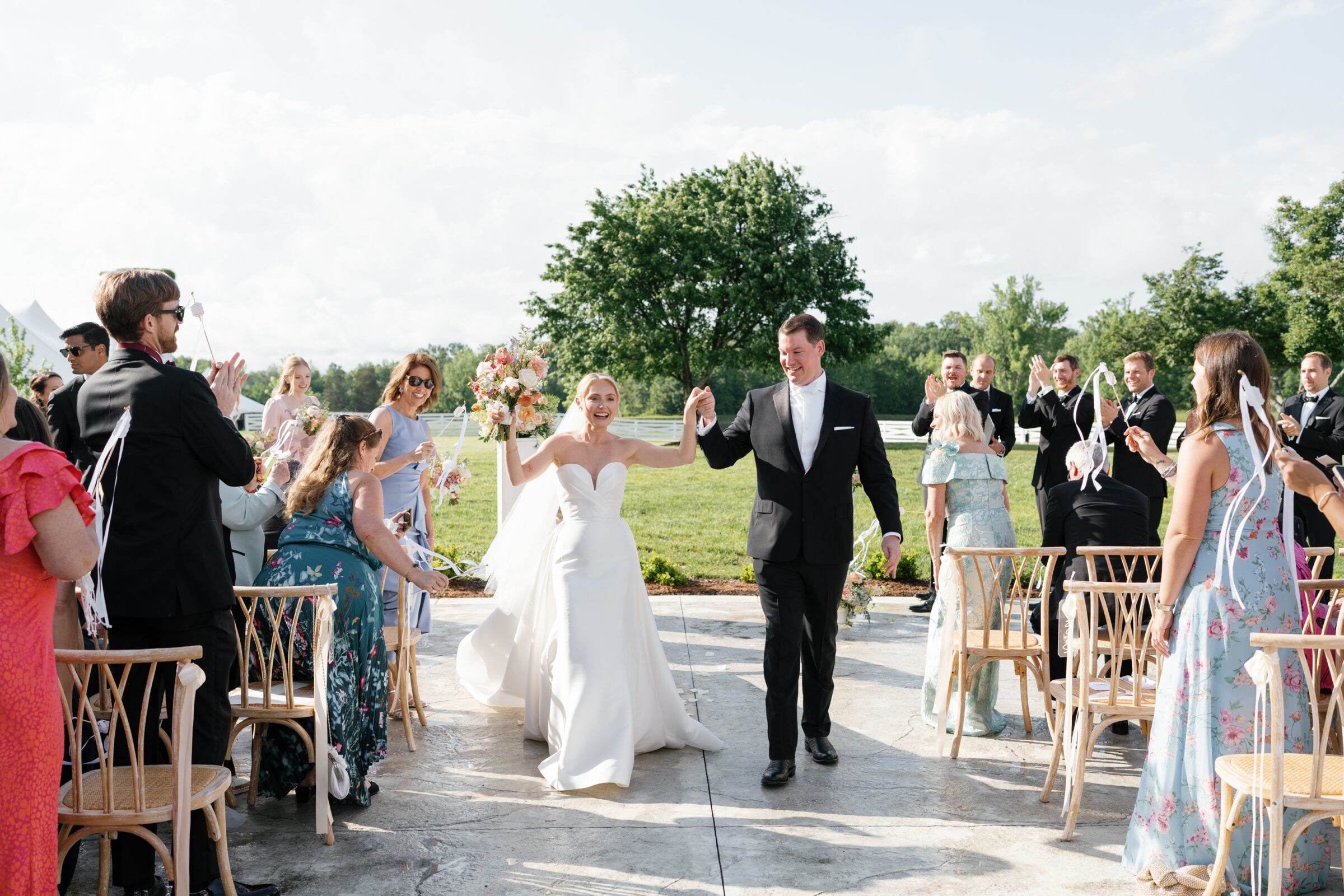 The joyous recessional: the newlyweds stride hand‑in‑hand down the aisle, beaming broadly. The bride raises her bouquet of peach, blush, and white blooms, while guests on either side wave ribbon wands in celebration. A single oak tree and bright green lawn form the backdrop beneath a clear sky.