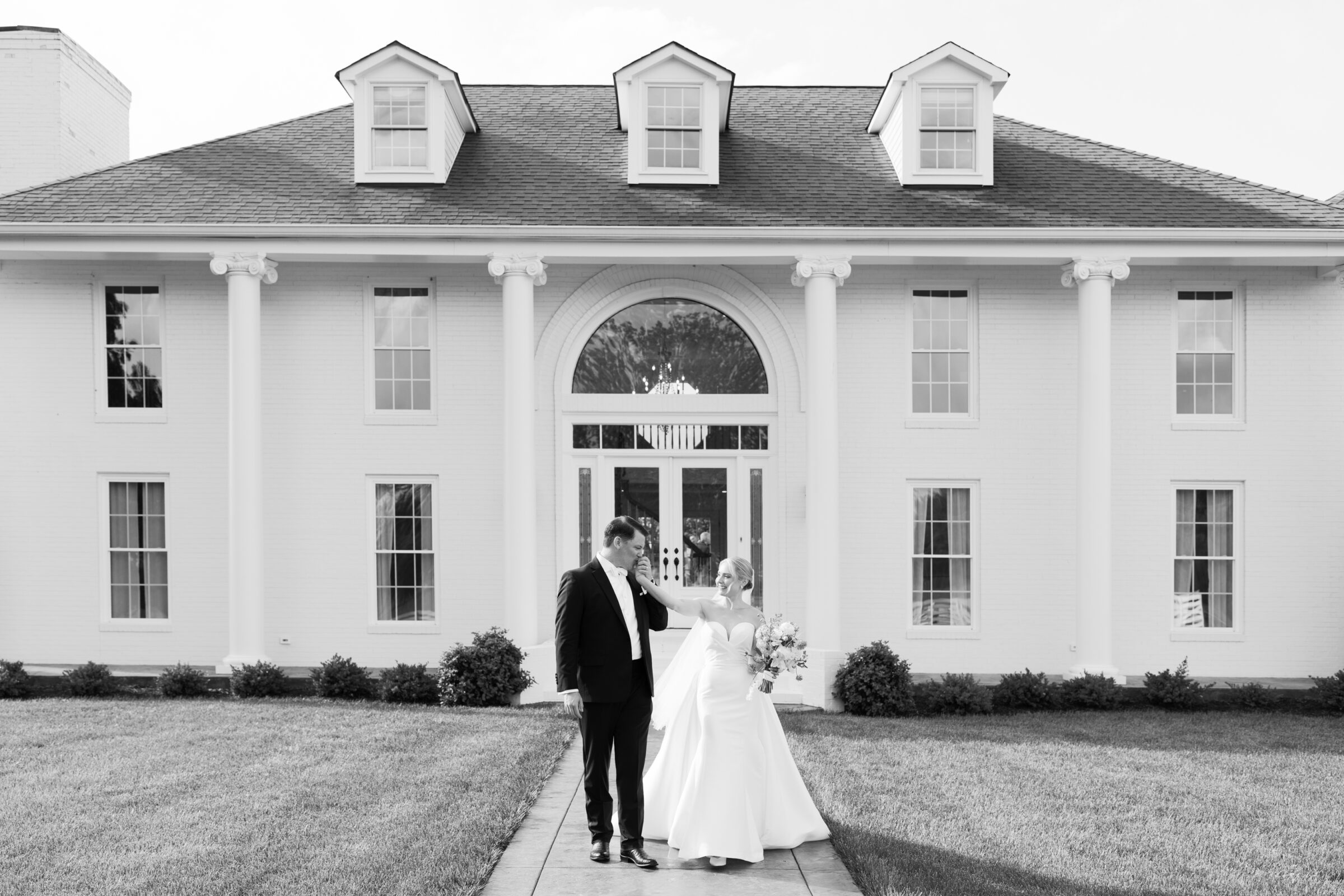 Northfield Estate Wedding - Richmond VA | A black-and-white shot of the newlyweds standing on a narrow walkway leading to the grand white-pillared facade of an estate. The bride, in a strapless gown and holding her bouquet, reaches out to offer her hand. The groom, in a dark tuxedo, gently takes and kisses it as they smile at each other beneath the three dormer windows and arched entryway.
