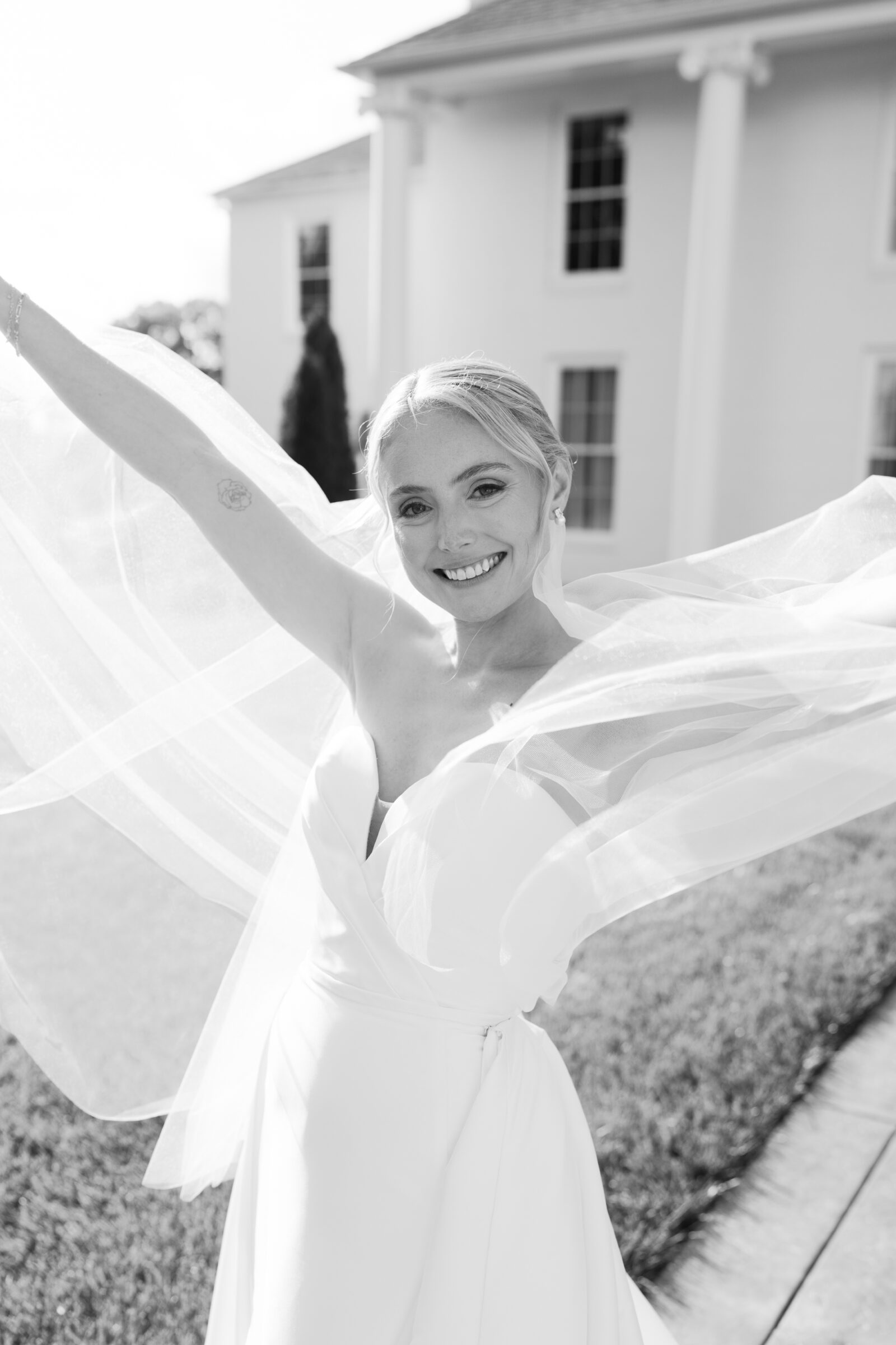 A black-and-white portrait of a smiling bride outdoors in front of a stately white-columned mansion. She lifts her arms, causing her long, sheer veil to billow around her like wings. She wears a strapless wedding gown and her hair is swept back in a low bun, and her joyful expression fills the frame.
