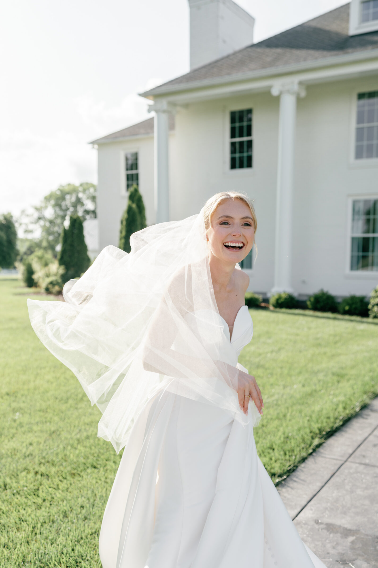 ChatGPT said:
A color portrait of a joyful bride standing on a slate walkway beside a manicured lawn, with a white-columned estate in the background. She wears a strapless white gown and a flowing veil that’s caught in the breeze, framing her laughing face. Her left hand gathers her veil as she leans forward, eyes bright and mouth open in delight.