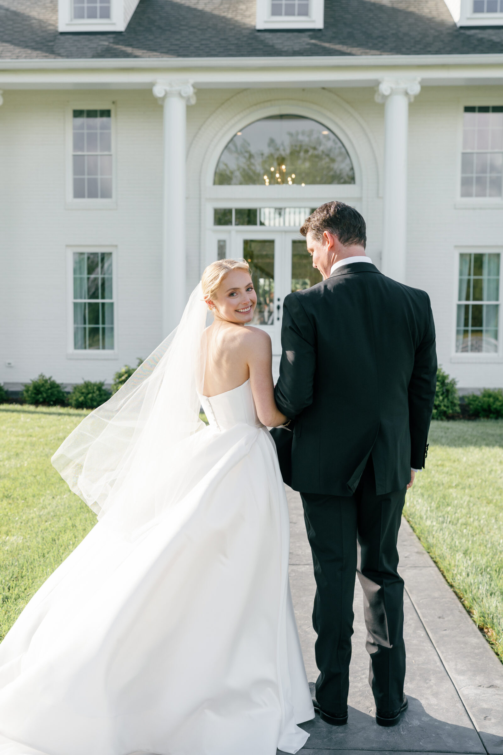 Northfield Estate Wedding - Richmond VA | A bride and groom walk arm‑in‑arm down a slate-gray path in front of a grand white-columned estate. The groom, in a black tuxedo, looks down at the path while the bride, in a strapless white gown and flowing veil, turns her head back over her shoulder and smiles at the camera.