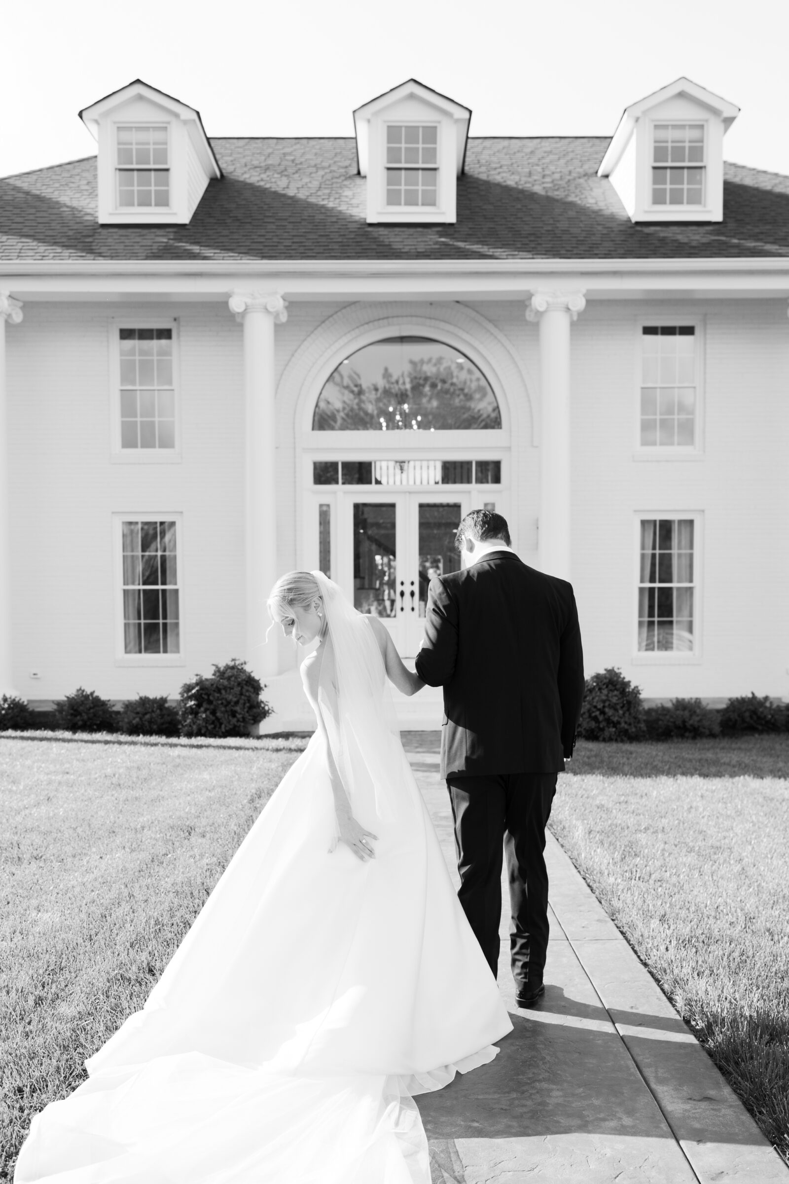 Northfield Estate Wedding - Richmond VA | A black‑and‑white rear‑view of the newlyweds as they stroll hand‑in‑hand along the slate pathway in front of the grand white manor. The groom leads, while the bride glances back over her shoulder, her sweeping train and long veil glowing in the sunlight beneath three dormer windows.