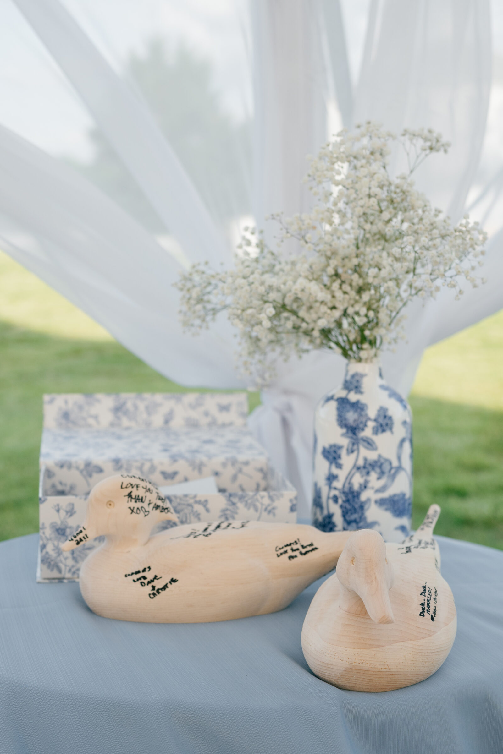 A round table draped in soft blue linen holds two unpainted wooden duck carvings covered in handwritten guest signatures, beside a matching blue-and-white porcelain vase brimming with delicate white baby’s breath and a gift box wrapped in coordinating blue toile paper, all set against billowing white drapery with sunlit grass beyond.