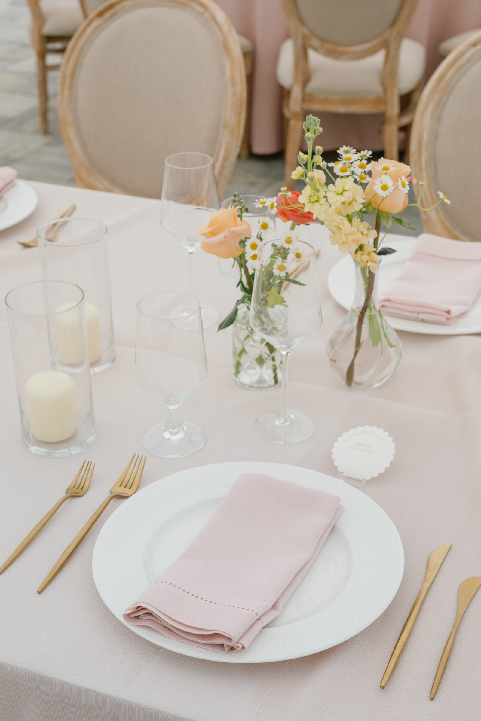 A close‐up of a softly lit place setting on a blush‑pink linen. In the foreground, a white charger holds a neatly folded pale rose napkin, flanked by slim gold forks on the left and matching knife and spoon on the right. Two clear glass cylinders hold ivory pillar candles, and three delicate vases of wildflower‑style blooms—small daisies, buttery yellow stock, and apricot‑toned roses—cluster behind the glassware. Two empty wine glasses stand ready, while a scalloped‑edge place card without any names peeks out from the background next to the flowers.
