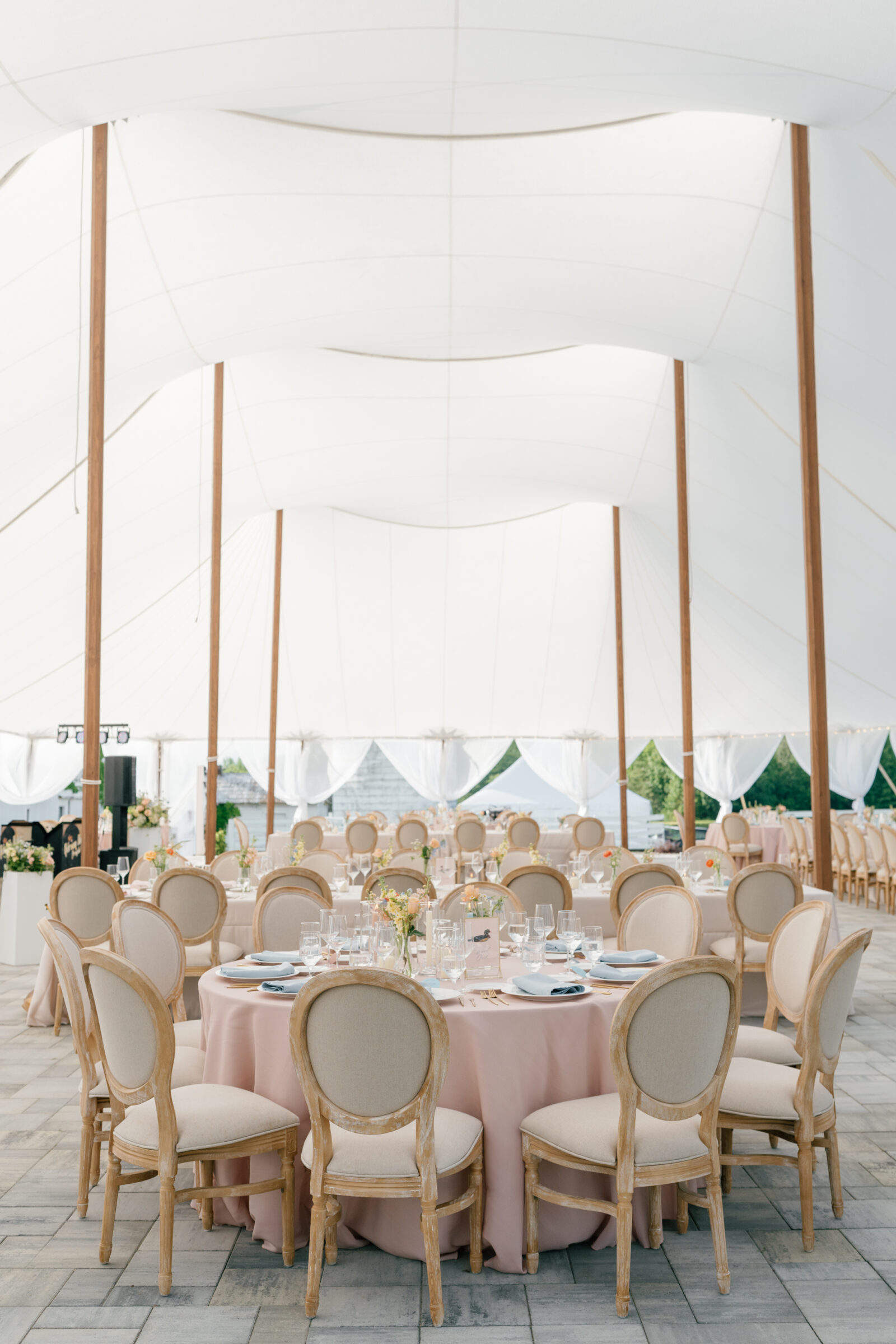 Northfield Estate Wedding - Richmond VA | Under a billowing white stretch tent held up by tall, slender wooden poles, round tables draped in soft blush linens are set for dinner. Each table is arranged with pale blue cloth napkins atop white plates, gold flatware, clear glassware, and small bouquets of pastel flowers. Light‑wood oval‑back chairs encircle the tables on a stone‑paved floor, while translucent drapes let in gentle daylight, creating an airy, elegant reception space.