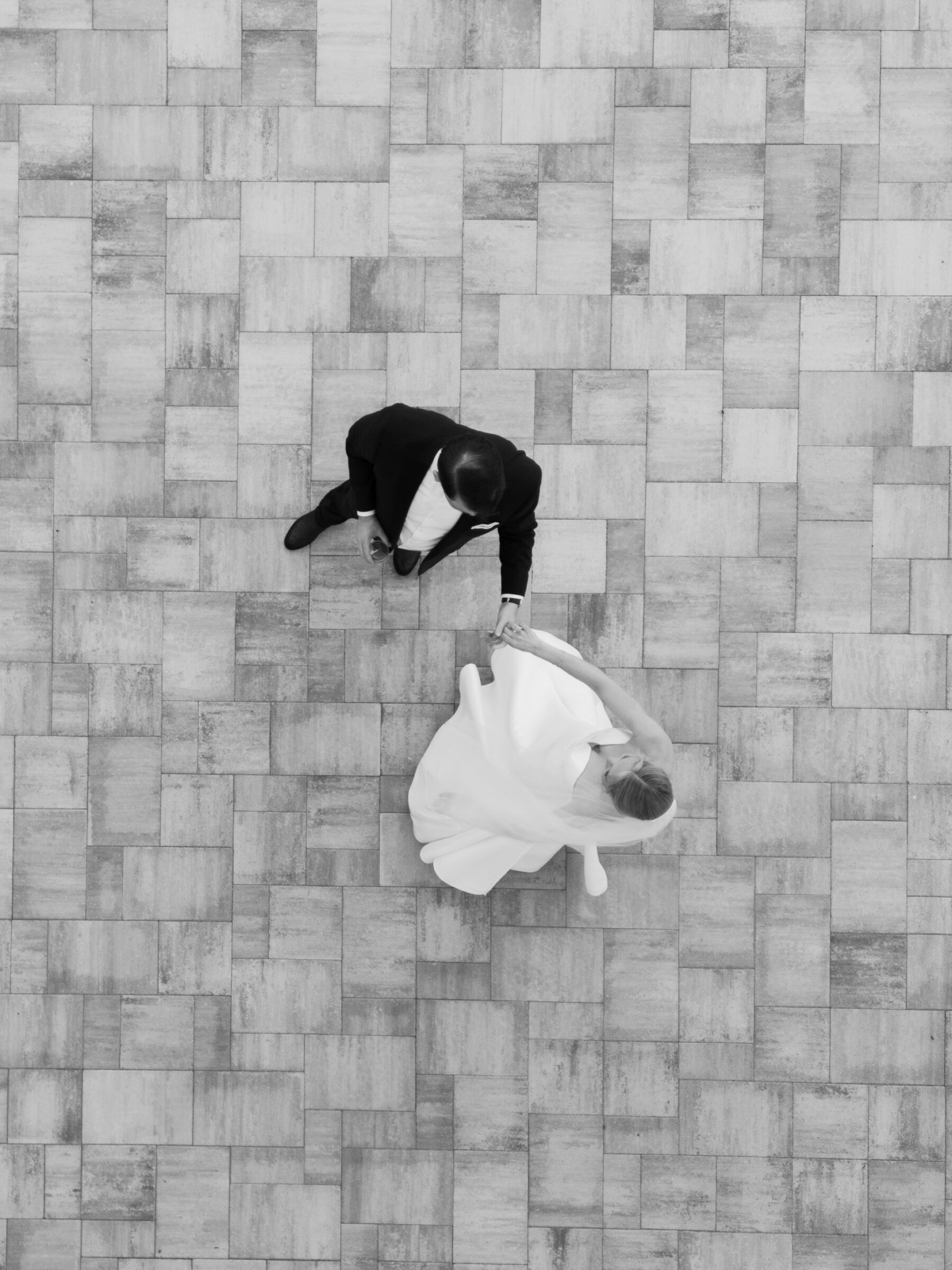 A black-and-white, top-down view shows the bride and groom holding hands as they move across a large, rectangular-tiled floor. The groom, dressed in a dark suit, leads with one arm extended, while the bride, in a flowing white gown and veil, follows beside him, their joined hands creating a gentle connection against the geometric pattern of the paving stones.