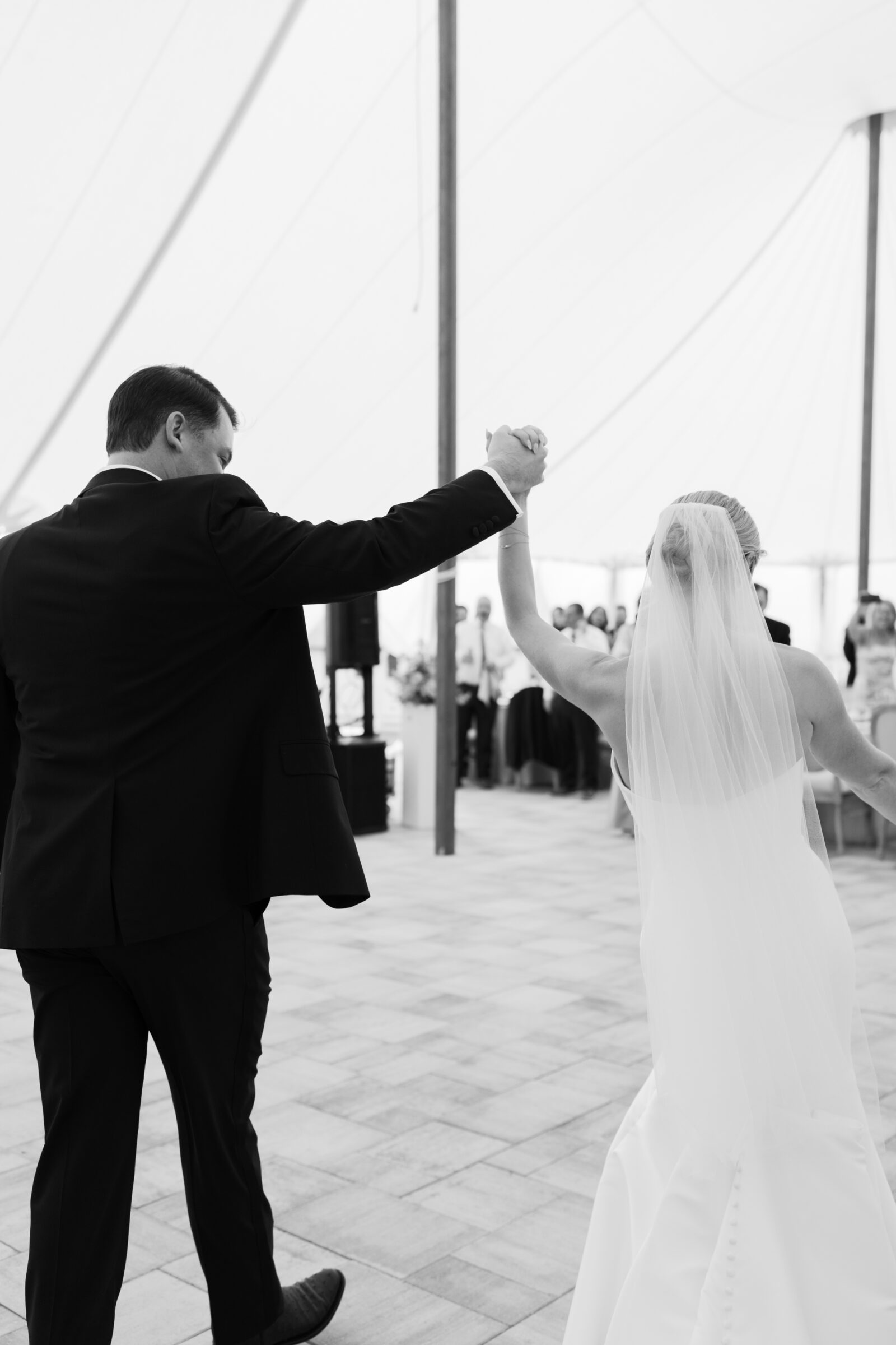A black-and-white image taken from behind shows the bride and groom stepping hand‑in‑hand under a white canopy into their reception. The groom wears a dark suit and the bride’s veil trails down her back as they enter the tented space, its fabric ceiling supported by tall wooden poles, with guests and ambient light visible just beyond them.