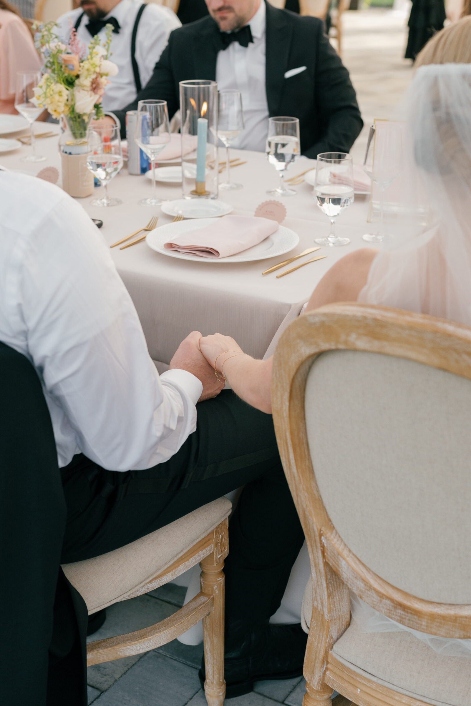A close-up color photo of a bride and groom seated side by side at a reception table, holding hands in their laps. The groom, in black tuxedo trousers and a white shirt, clasps the bride’s hand, which rests on her flowing white wedding gown. In front of them, a neatly set place setting features a white plate topped with a folded blush‑pink napkin, flanked by gold flatware, clear stemware, and a single lit taper candle in a glass cylinder. Elegant wooden‑frame chairs and a soft stone‑tile floor complete the scene.