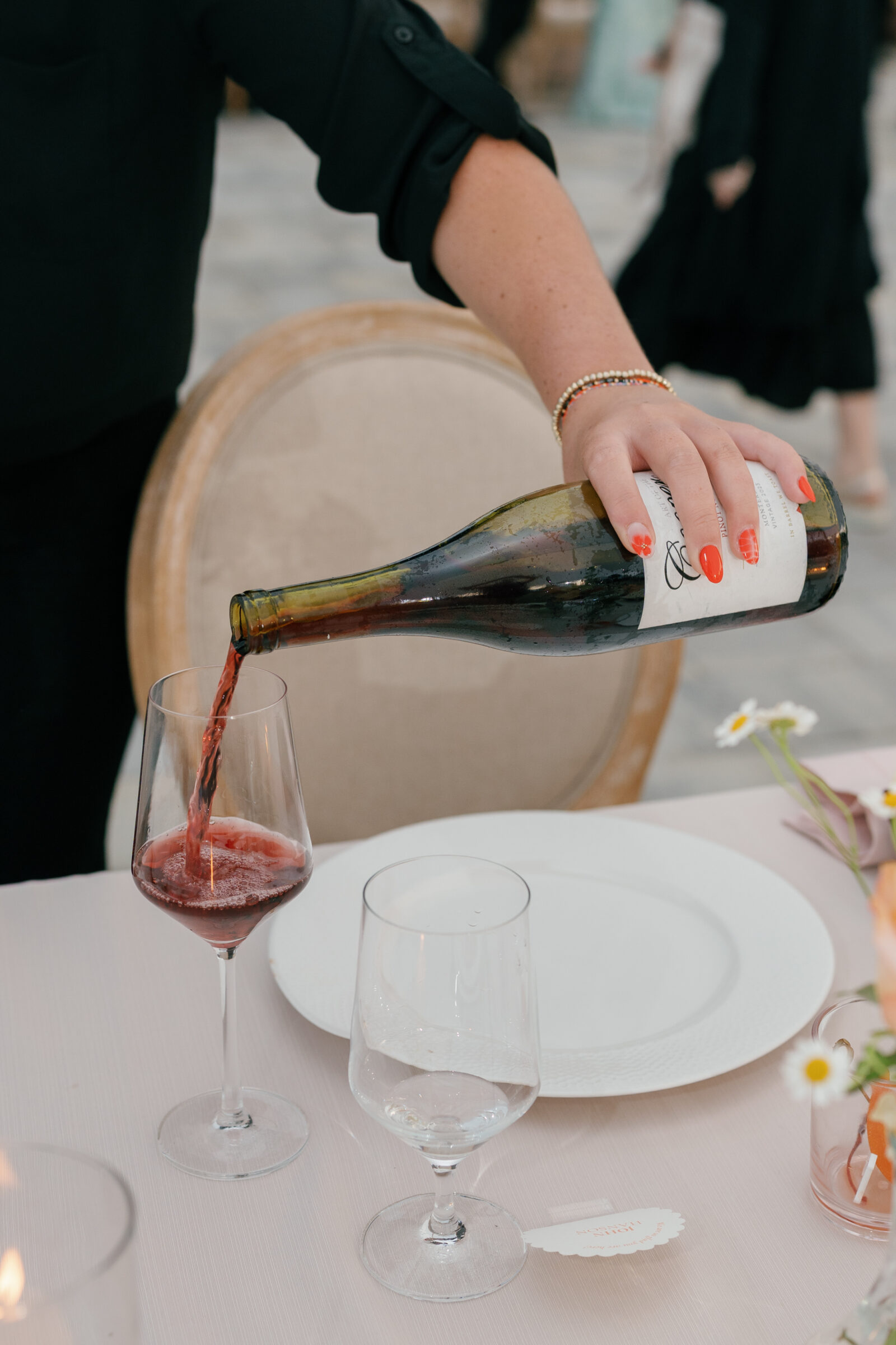 A close-up color photo showing a server’s hand pouring red wine from a dark green bottle into a tall, slender wine glass. The hand, wearing bright orange-red painted nails and a delicate beaded bracelet, tilts the bottle so a stream of deep ruby liquid swirls into the glass. In the foreground, a second smaller water glass stands empty beside a white dinner plate on a blush‑pink tablecloth. Soft candlelight and out‑of‑focus floral blooms peek in at the edges of the frame.