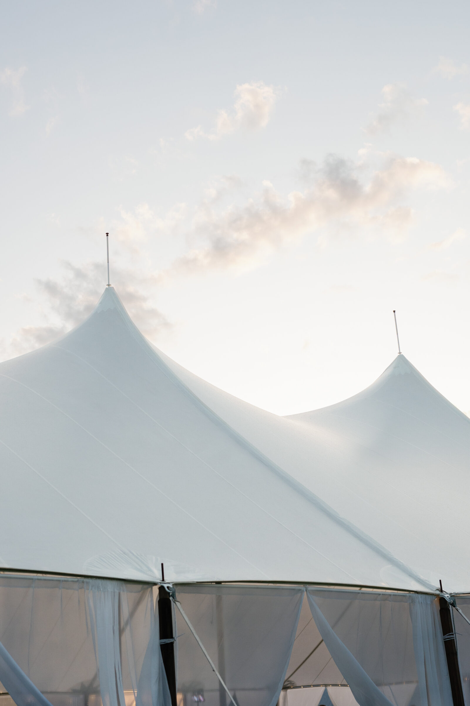 A close-up of two white sailcloth tent peaks rising against a pale evening sky. Each pointed apex is topped with a slender finial, and soft ripples in the taut fabric catch the fading sunlight. The tent’s translucent side walls and draped panels are visible below, hinting at the reception space inside.