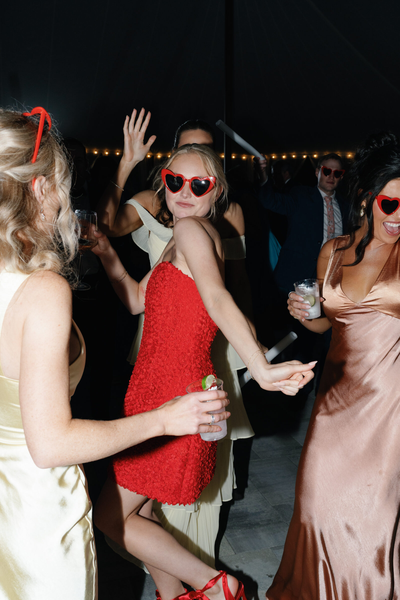 Bride in a red textured mini dress and heart-shaped sunglasses dances with guests during a lively spring wedding reception at Northfield Estate in Cumberland, VA, photographed by a Richmond wedding photographer using direct flash for an editorial party vibe.