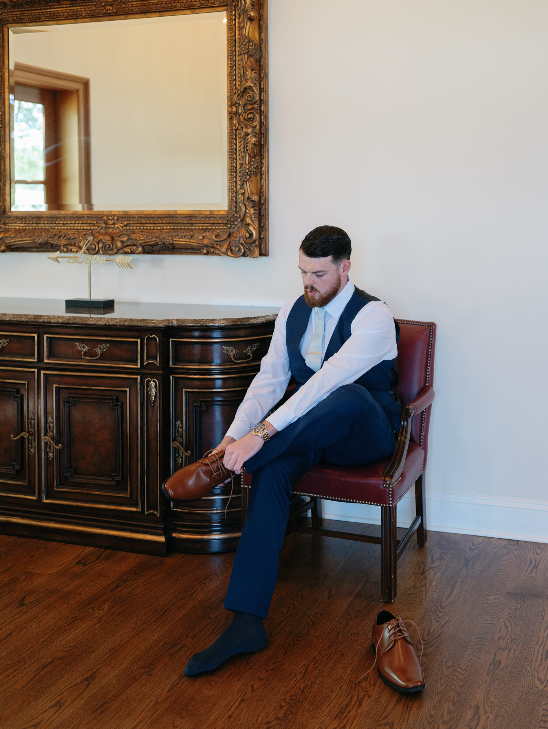 Groom in navy suit and pale blue tie sits in a refined Trump Winery Wedding preparation room in Charlottesville, lacing his brown leather shoes beneath an ornate gold‑framed mirror