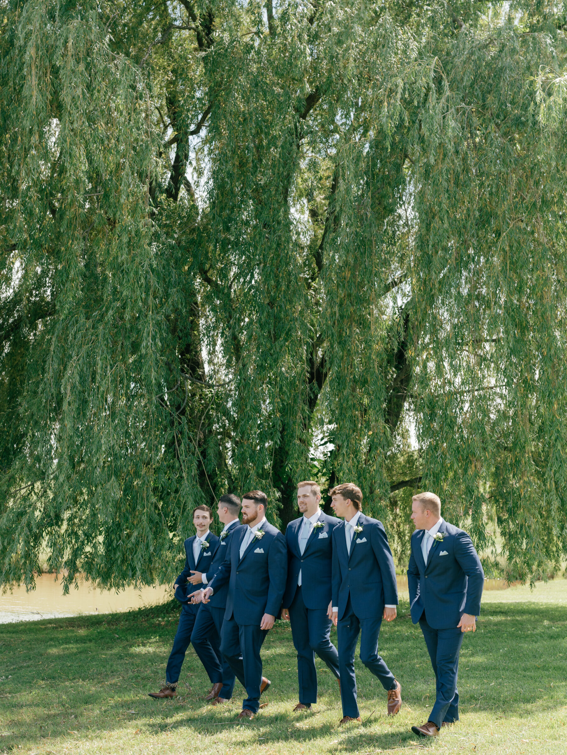 Groomsmen in matching navy suits strolling beneath a sweeping willow tree on the manicured grounds of a Trump Winery Wedding in Charlottesville