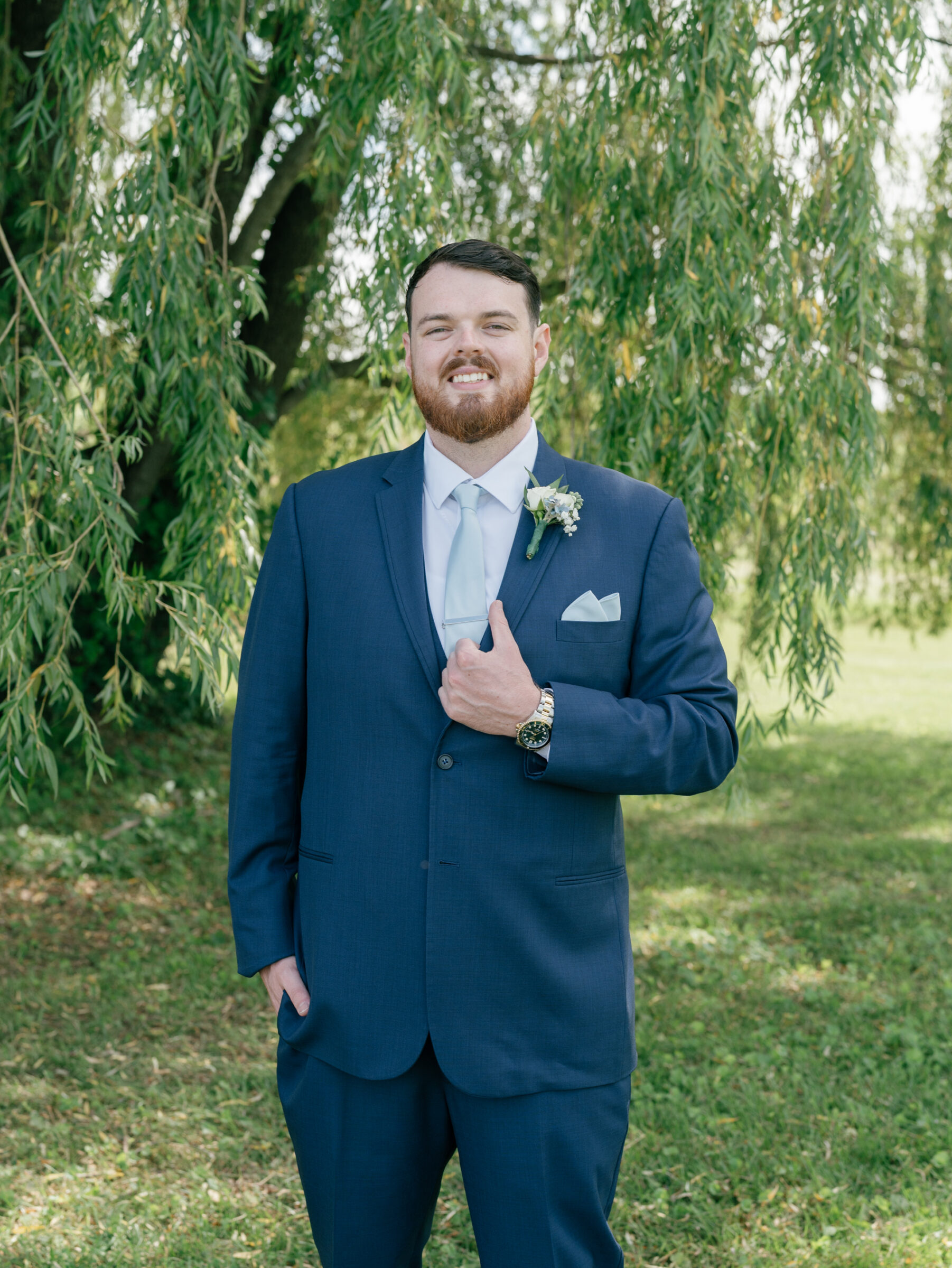 Close‑up portrait of smiling groom in navy suit, pale blue tie, and boutonniere standing beneath a cascading willow at a Trump Winery Wedding in Charlottesville