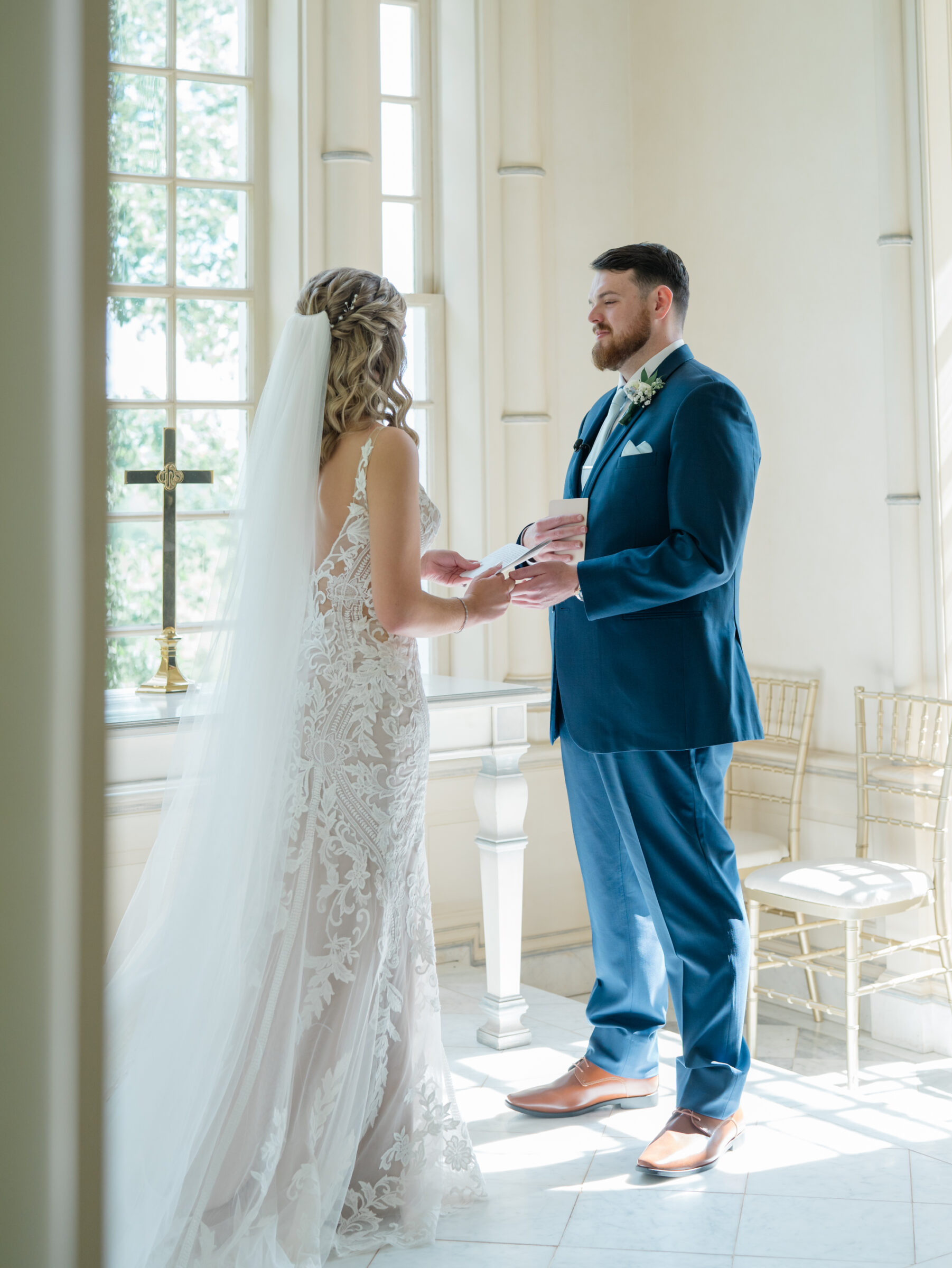 Bride in intricate lace gown and groom in navy suit exchange heartfelt vows beside a sunlit window at their Trump Winery Wedding in Charlottesville