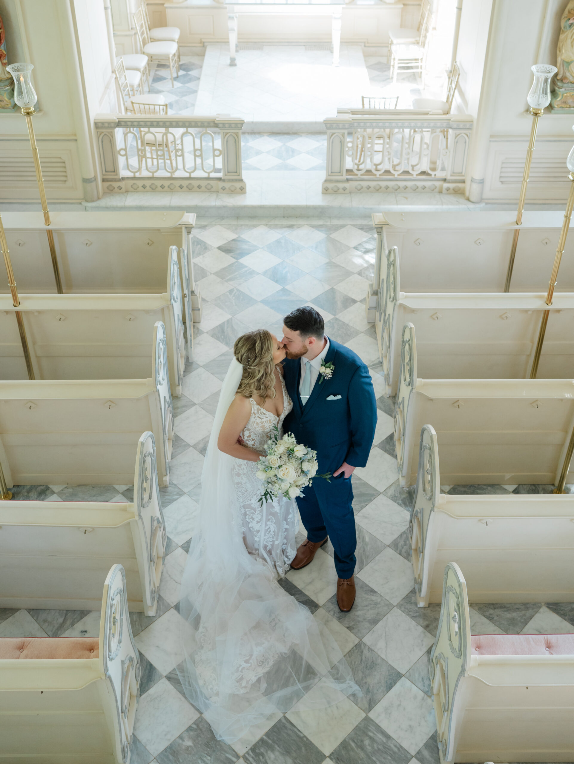 Aerial view of bride in lace gown and groom in navy suit sharing a kiss in the marble‑tiled aisle of a Trump Winery Wedding chapel in Charlottesville