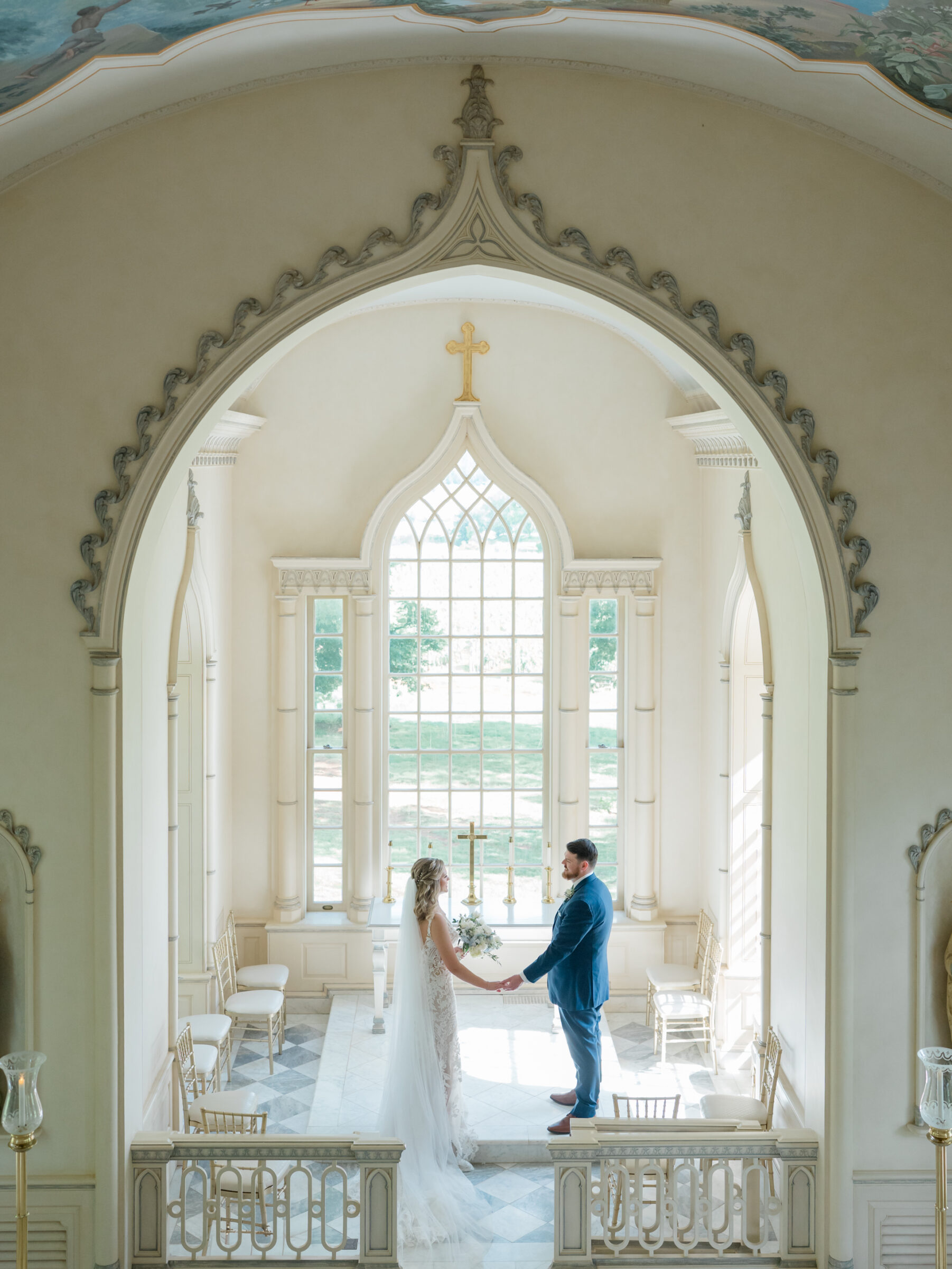 Bride in a lace gown and groom in a navy suit holding hands at a sunlit altar beneath an ornate vaulted arch during their Trump Winery Wedding in Charlottesville