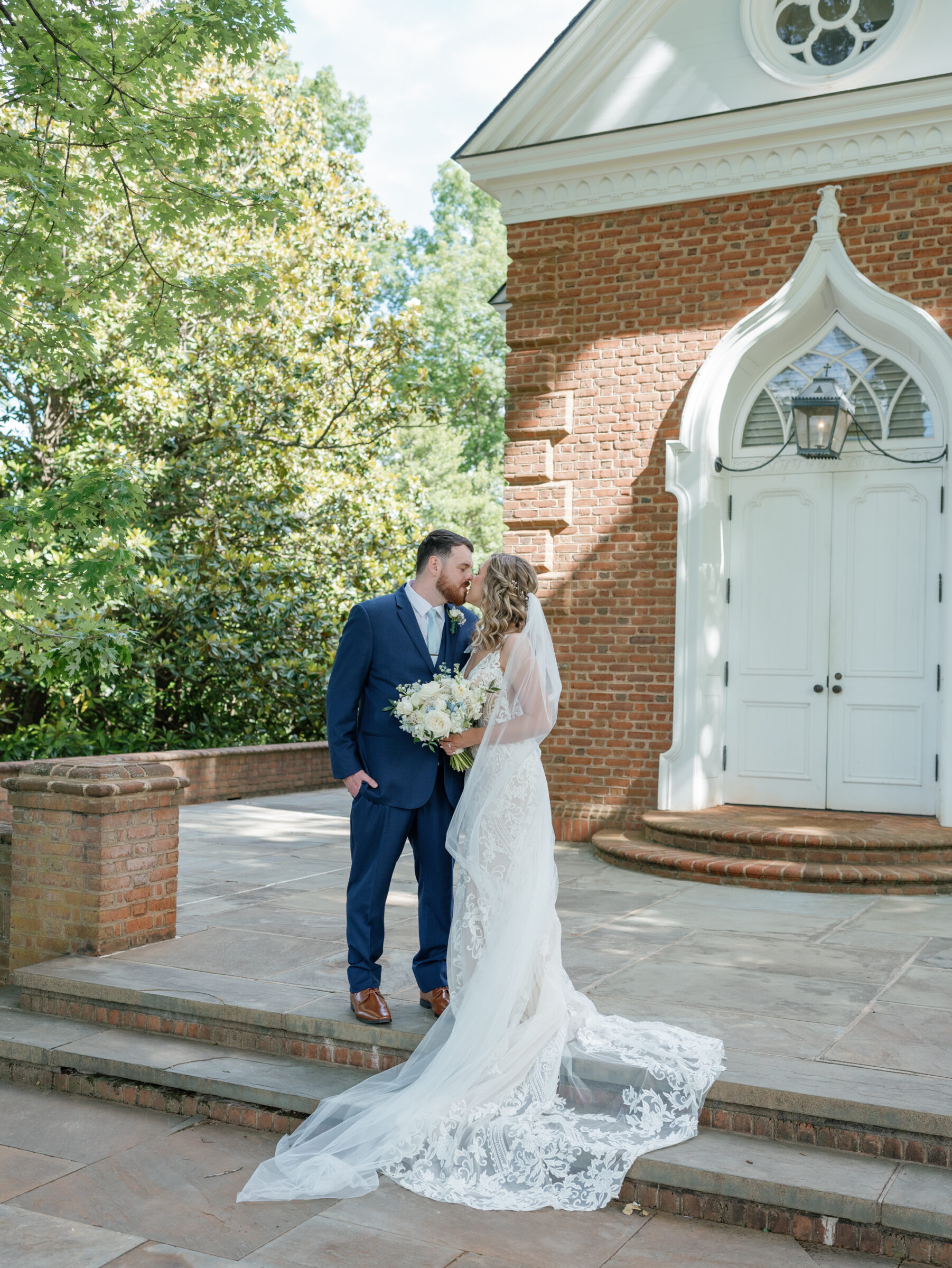 Bride in a lace gown with flowing train and veil kisses her groom in a navy suit on the brick chapel steps during their Trump Winery Wedding in Charlottesville