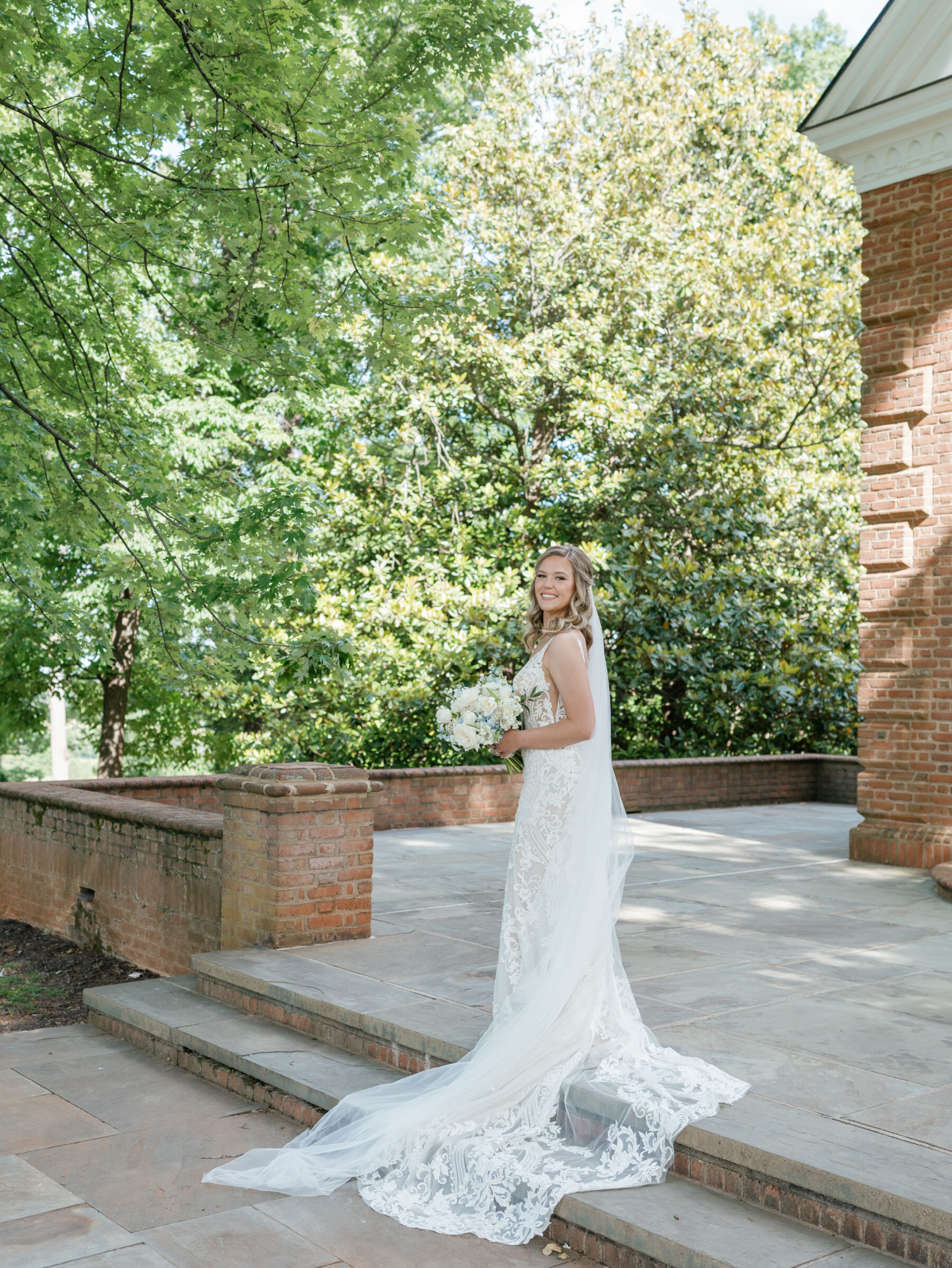 Smiling bride in a lace gown with a flowing train and veil holding a white-and-blue bouquet on the brick chapel terrace at a Trump Winery Wedding in Charlottesville