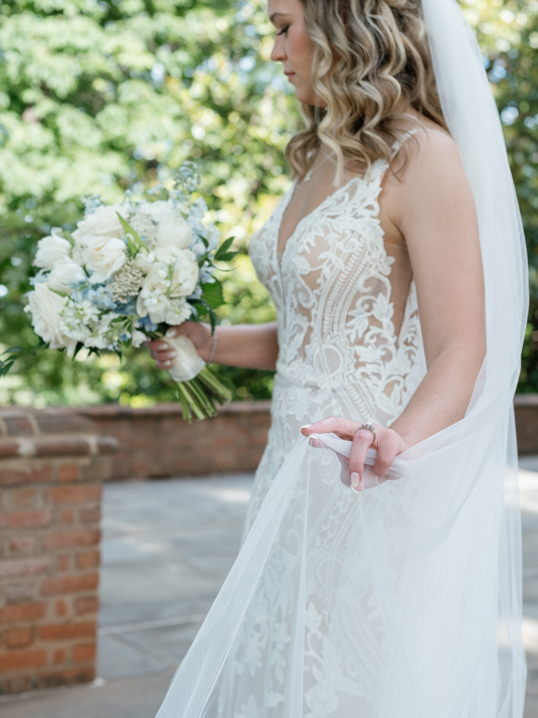 Detail of bride’s hand delicately lifting her tulle veil while holding a white-and-blue bouquet during a Trump Winery Wedding in Charlottesville