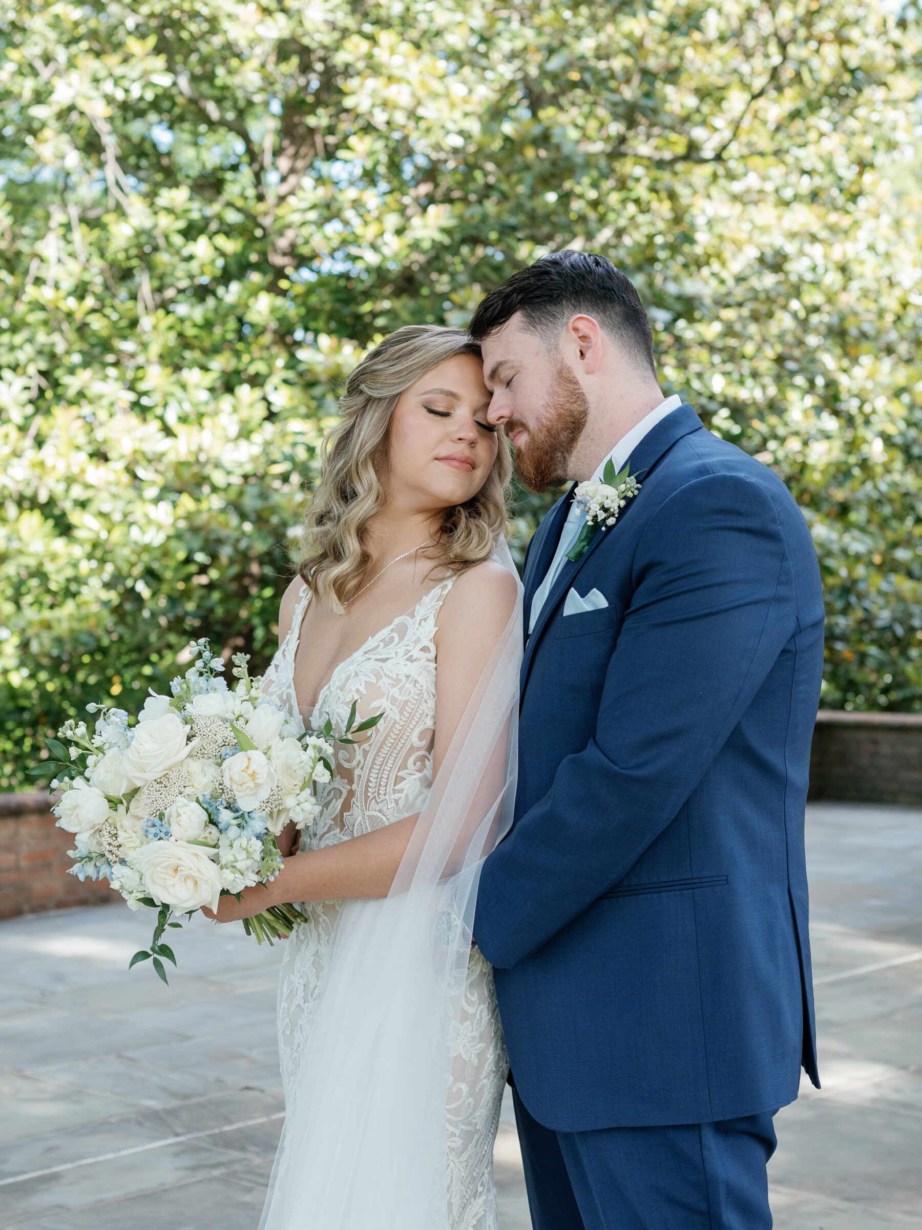 Bride in lace wedding gown holding a white-and-blue rose bouquet as her groom in a navy suit gently embraces her beneath lush greenery at a Trump Winery Wedding in Charlottesville