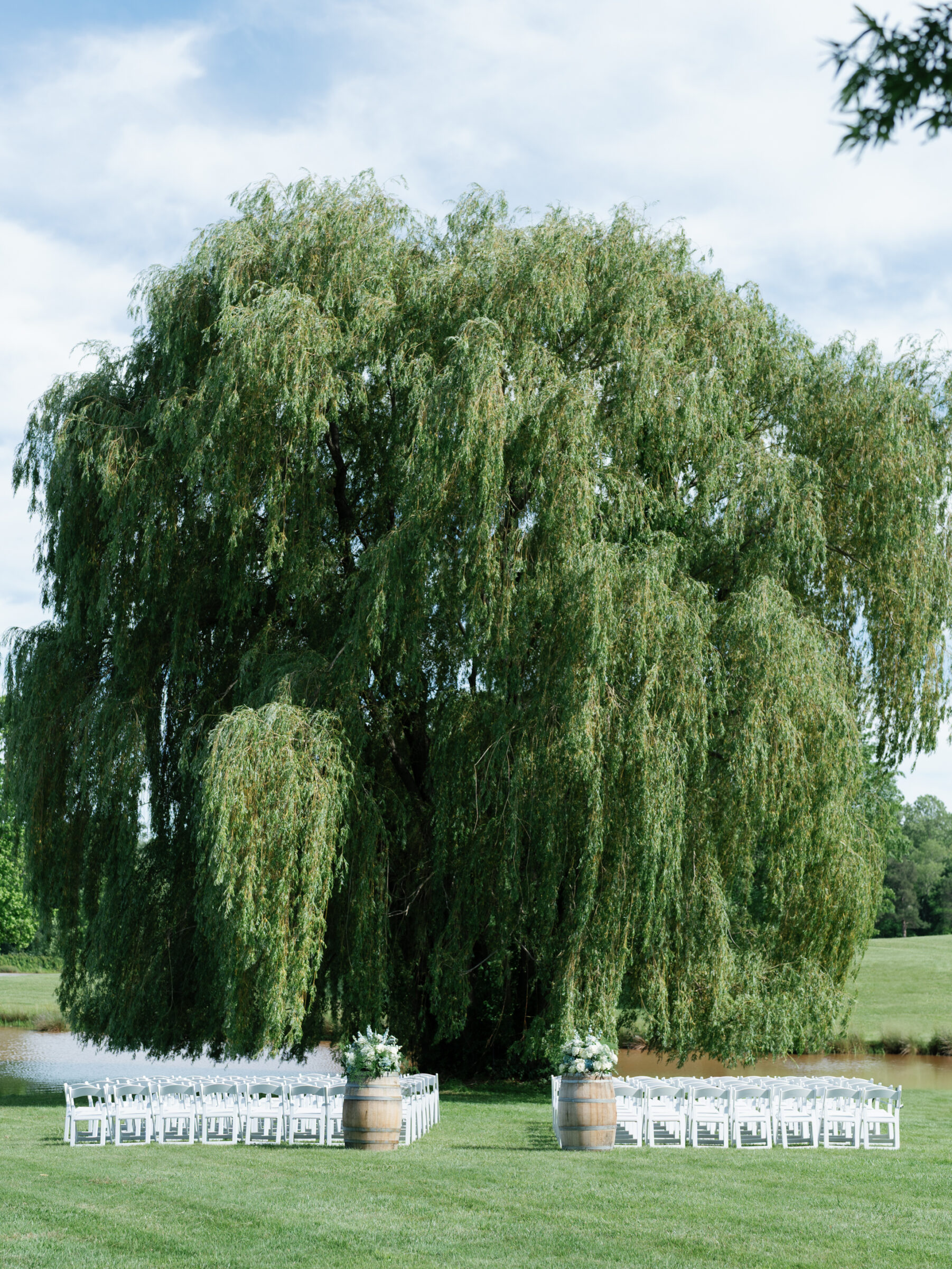 Rows of white folding chairs set between floral‑topped wine barrels beneath a sweeping willow tree by the pond at a Trump Winery Wedding in Charlottesville