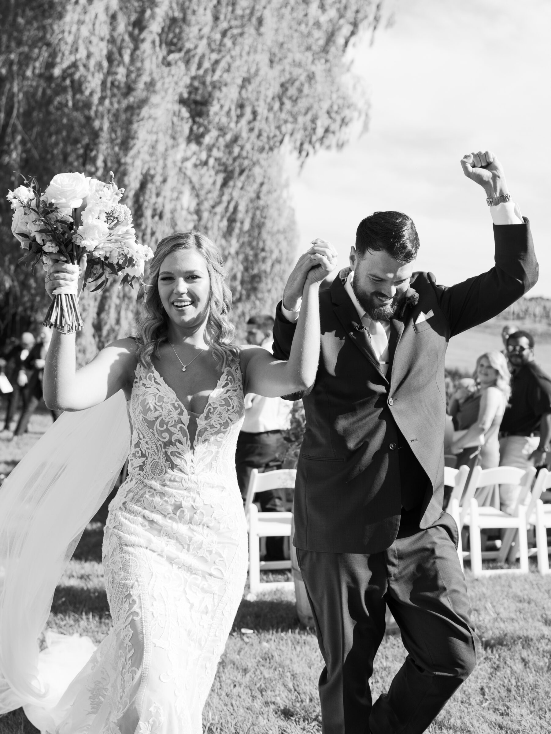 Black‑and‑white image of bride in lace gown and groom in tailored suit raising arms in celebration as they exit their Trump Winery Wedding ceremony under a willow