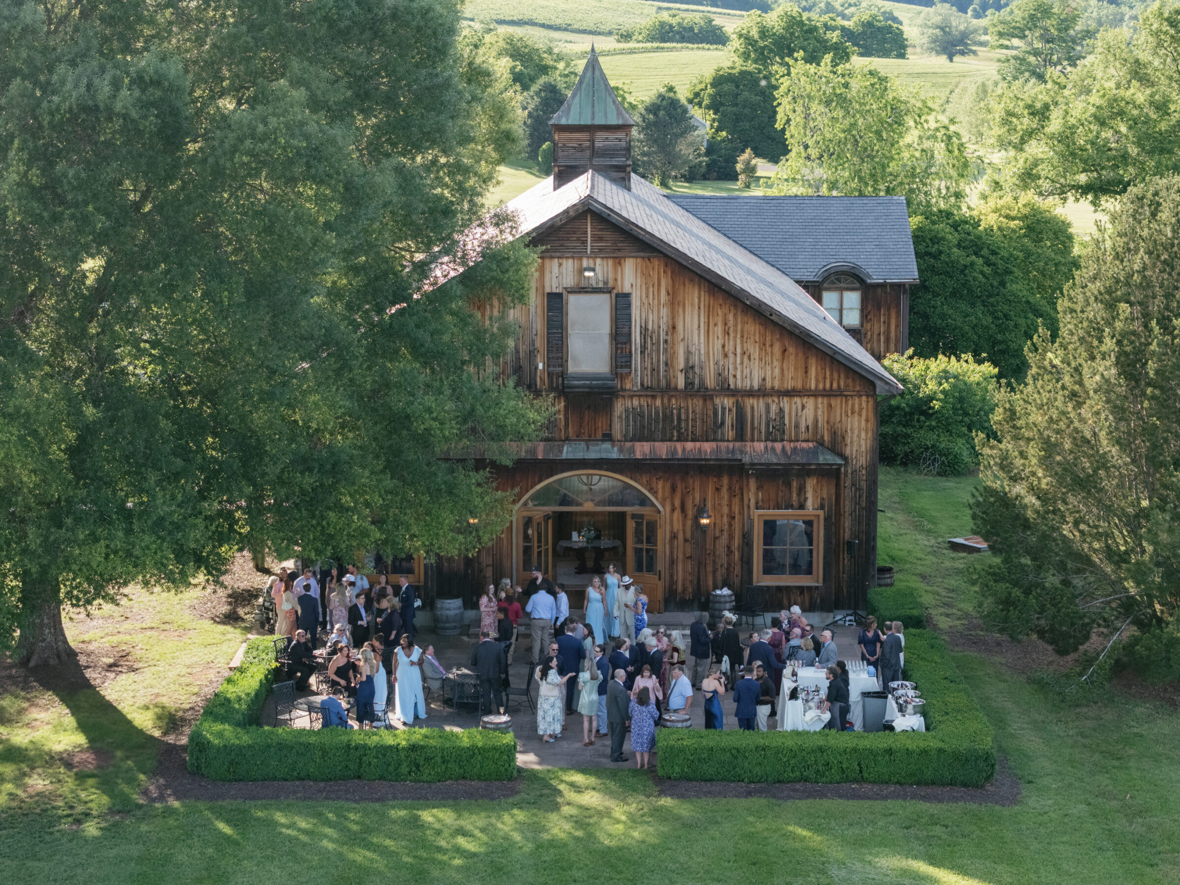 Wide-angle aerial view of guests mingling on the manicured terrace in front of the rustic barn reception venue at a Trump Winery Wedding in Charlottesville