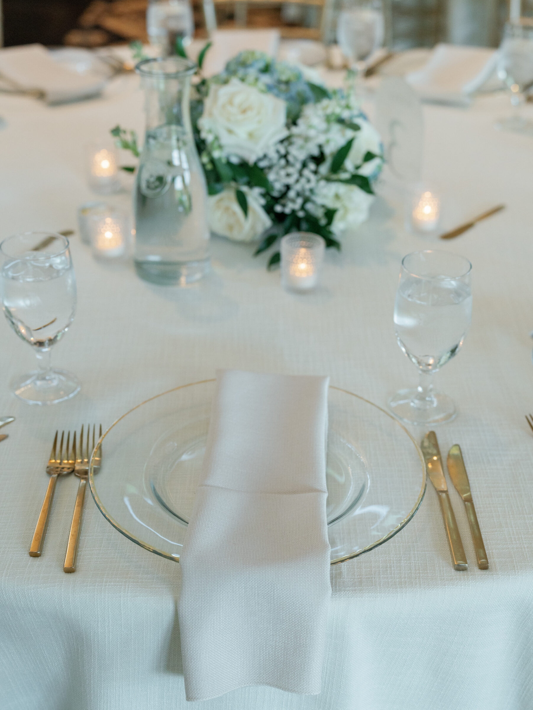 Close‑up of ivory‑draped table at a Trump Winery Wedding reception in Charlottesville, featuring a gold‑rimmed glass charger, soft taupe linen napkin, gleaming gold flatware, water goblets, and a white‑and‑blue rose centerpiece with votive candles