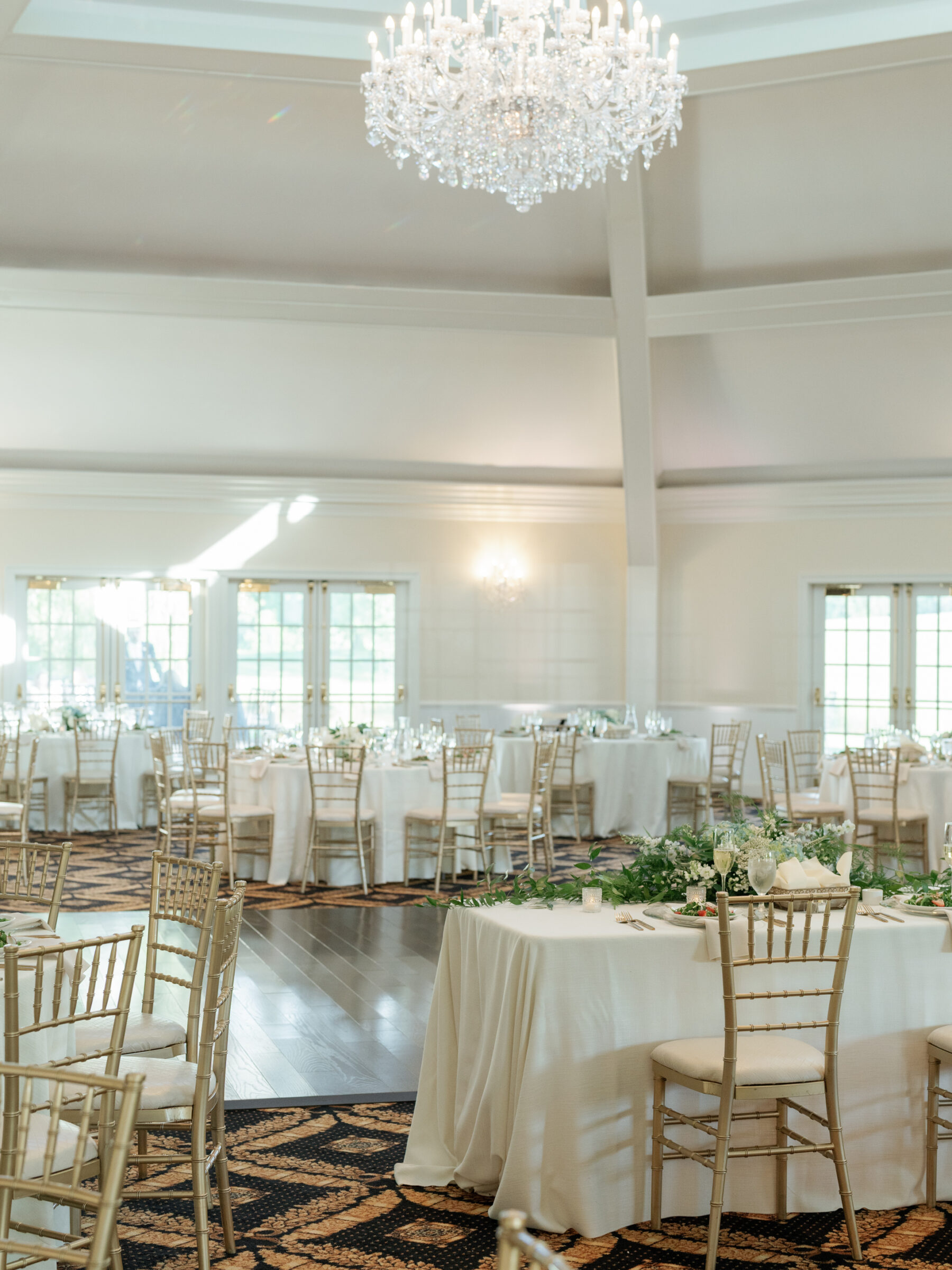Elegant reception ballroom at a Trump Winery Wedding in Charlottesville, featuring a sparkling crystal chandelier overhead, ivory‑draped tables with lush greenery runners, and gold Chiavari chairs arranged around the polished wood dance floor