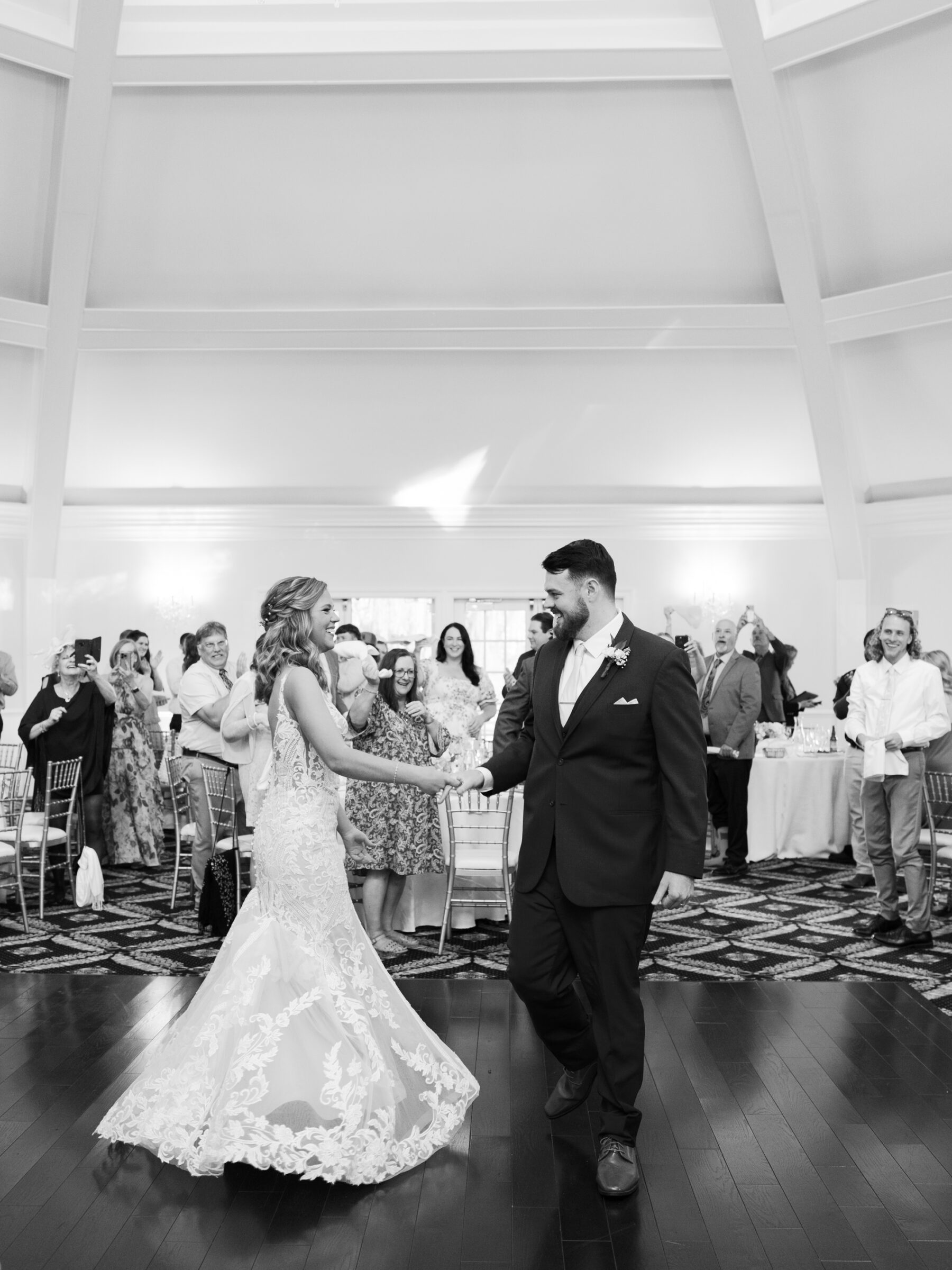 Black‑and‑white image of bride in lace mermaid gown and groom in tailored suit sharing their first dance on the polished wood floor beneath a vaulted ceiling at their Trump Winery Wedding reception in Charlottesville