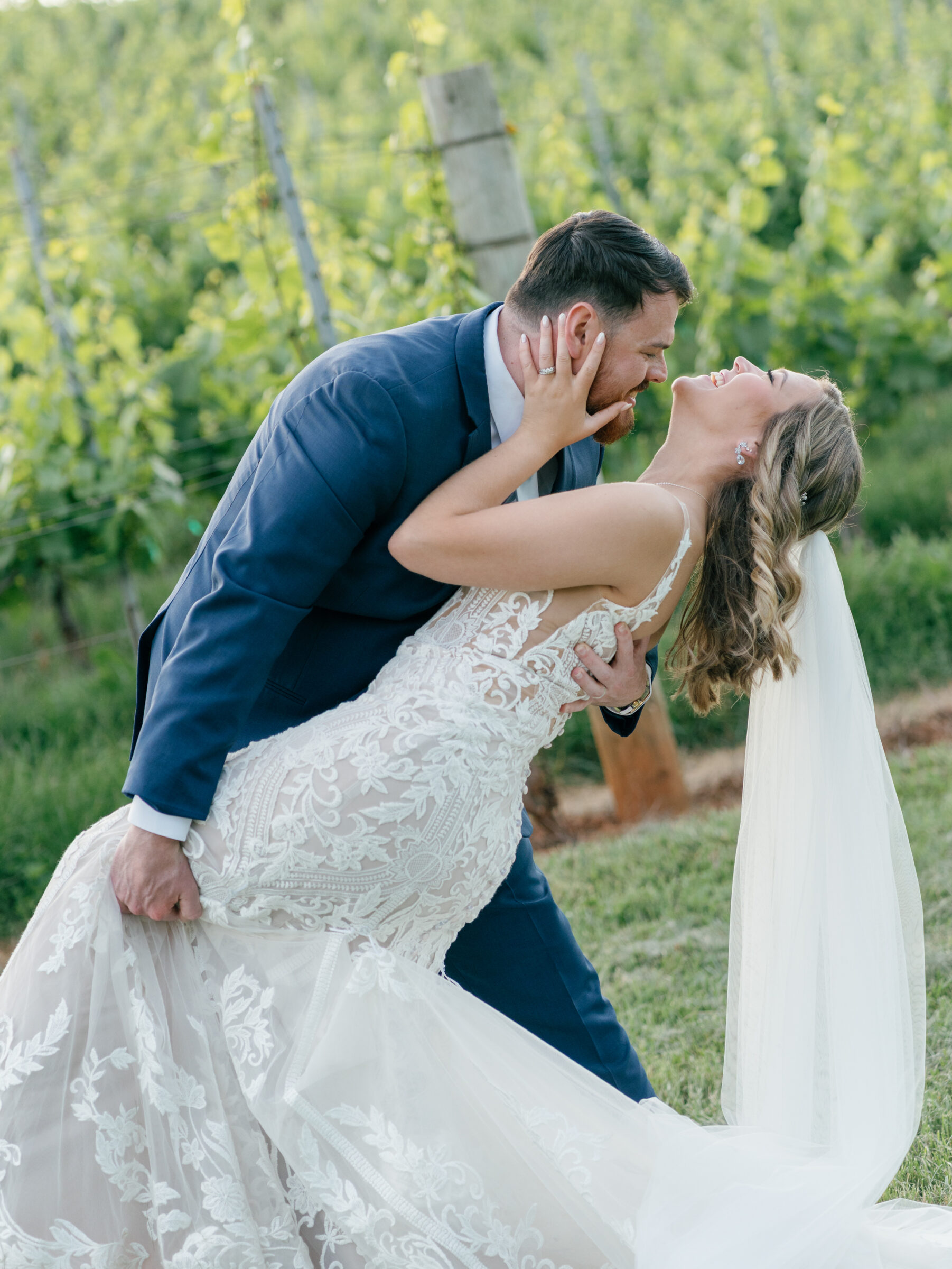 Groom in navy suit gently dips his bride in a lace mermaid gown and long tulle veil for a joyful kiss among the vineyard rows at a Trump Winery Wedding in Charlottesville