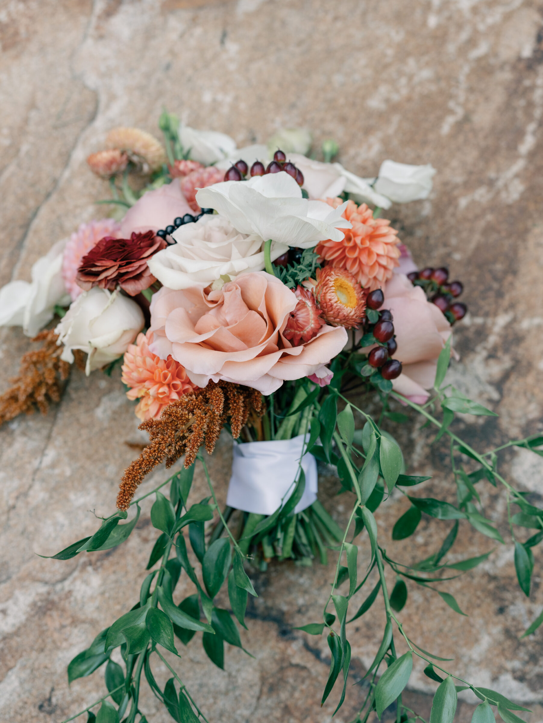 Bridal bouquet with peach roses, dahlias, anemones, and greenery resting on stone.
